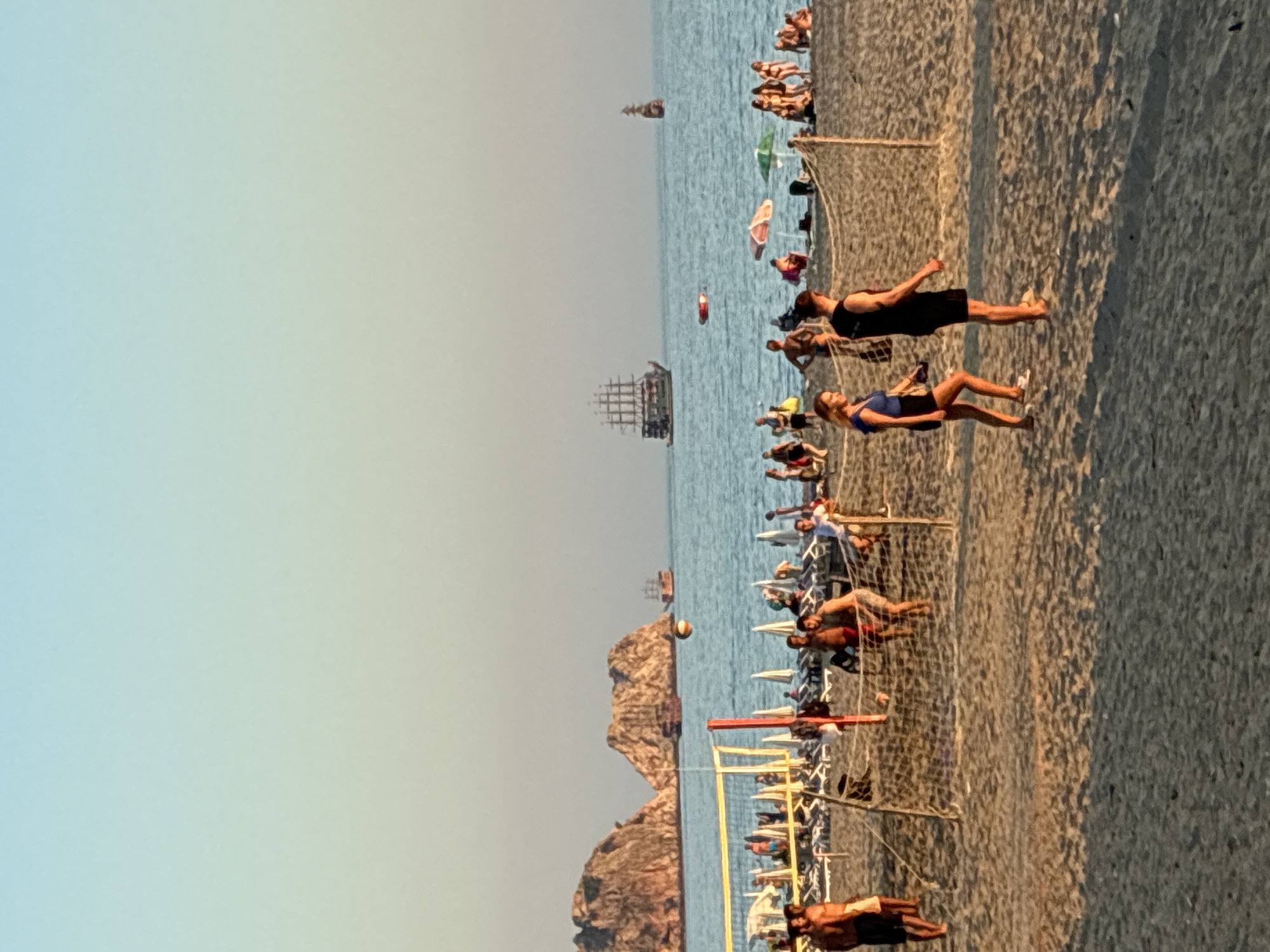 The image depicts a beach scene in Alanya, Turkey, with people engaged in recreational activities. In the foreground, a sandy beach hosts several individuals. A volleyball net is set up, and a group of men appear to be playing volleyball. Two individuals, a woman and a man, are walking on the beach in the foreground, in front of the volleyball net. Further back, more people are visible relaxing on lounge chairs under umbrellas, and others are walking along the shoreline.
The sea occupies the middle ground, with two large sailing ships, resembling pirate ships, in the distance. Smaller boats are also present on the water. The iconic Alanya Castle and its surrounding rock formations are visible on the left side of the image, jutting out into the sea, with a smaller rock formation to its right. The sky above is a clear gradient of light blue transitioning to a lighter hue near the horizon. The lighting suggests it is late afternoon or early evening, with the setting sun casting long shadows on the sand. The overall atmosphere is one of leisure and vacation.