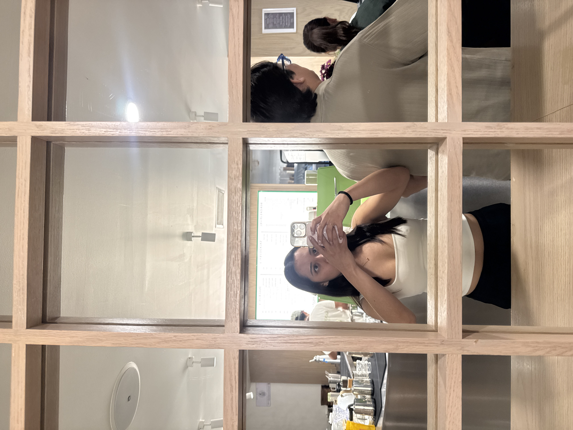 A woman in a white top and black pants is seen reflected in a window. She is holding a cell phone with two hands in front of her face as if taking a photo. The window is made up of a grid of light-colored wood. A blurry reflection of other people can be seen in the top pane of the window. The background of the reflection in the middle pane appears to be a cafe or restaurant with a menu board. Various objects, possibly coffee making equipment, are reflected in the bottom pane. The location appears to be in the city of Puebla, Mexico.