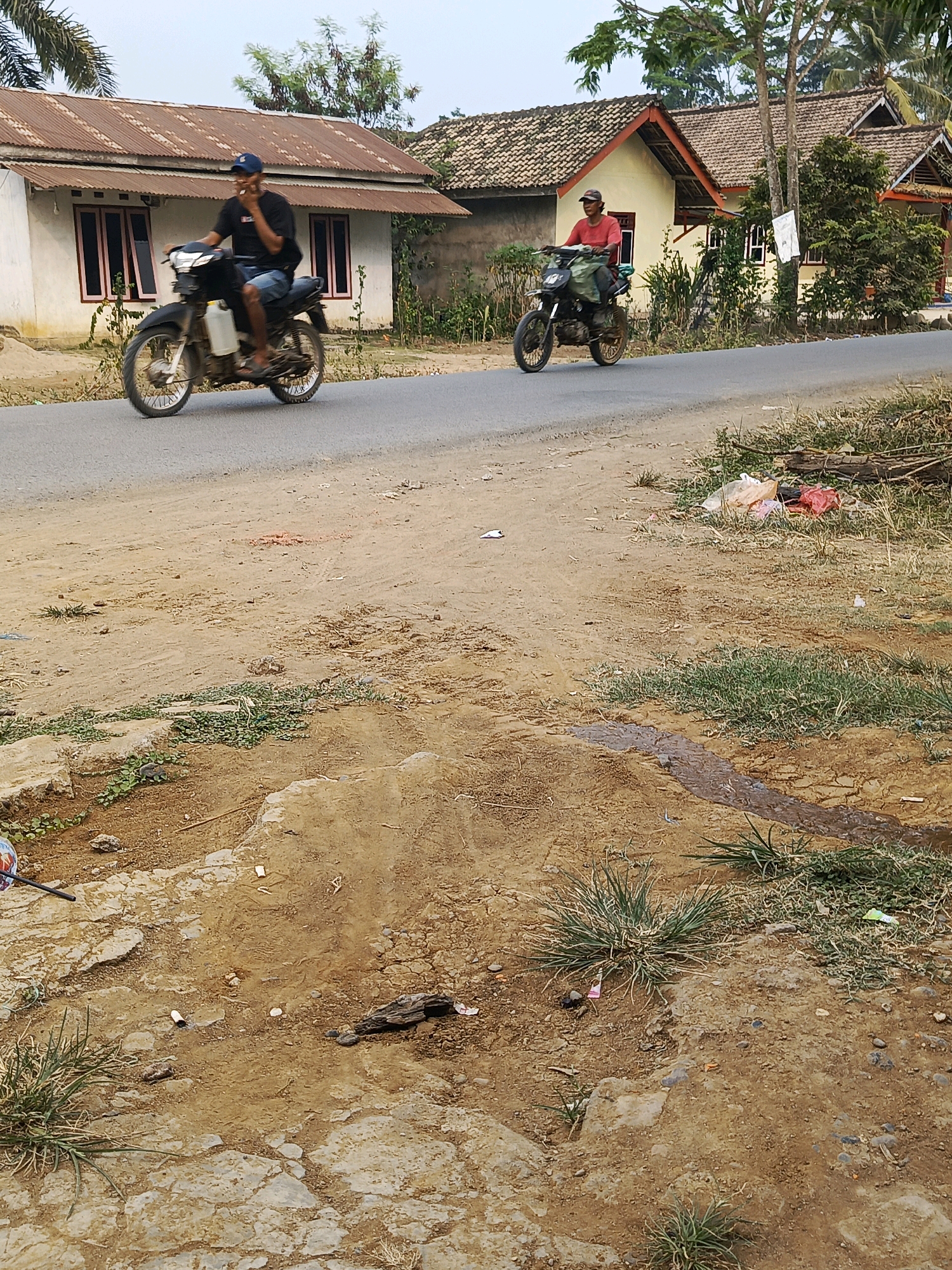 In Megang Sakti I, Indonesia, two men are riding motorcycles on a paved road. The man on the left, wearing a black t-shirt and blue shorts, is on a black motorcycle with a white gas tank. The man on the right, wearing a red t-shirt, is on a similar black motorcycle. They are riding past white houses with corrugated metal roofs and brown roof tiles. The road is bordered by a dirt shoulder with scattered grass, debris, and a small puddle of water. The environment appears to be rural or semi-rural.