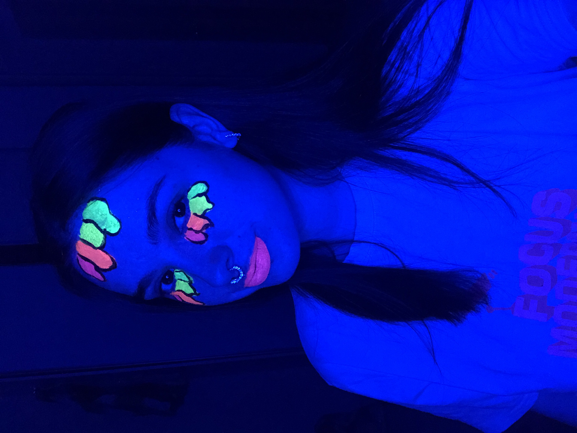 A woman with dark hair lies on her side under a blacklight. She has fluorescent face paint in green and orange around her eyes and on her forehead, and fluorescent pink on her lips. She has a ring in her nostril and an earring. She is wearing a white shirt with the text "FOCUS MODEN" visible on the front. The background is dark. The location is not discernible.