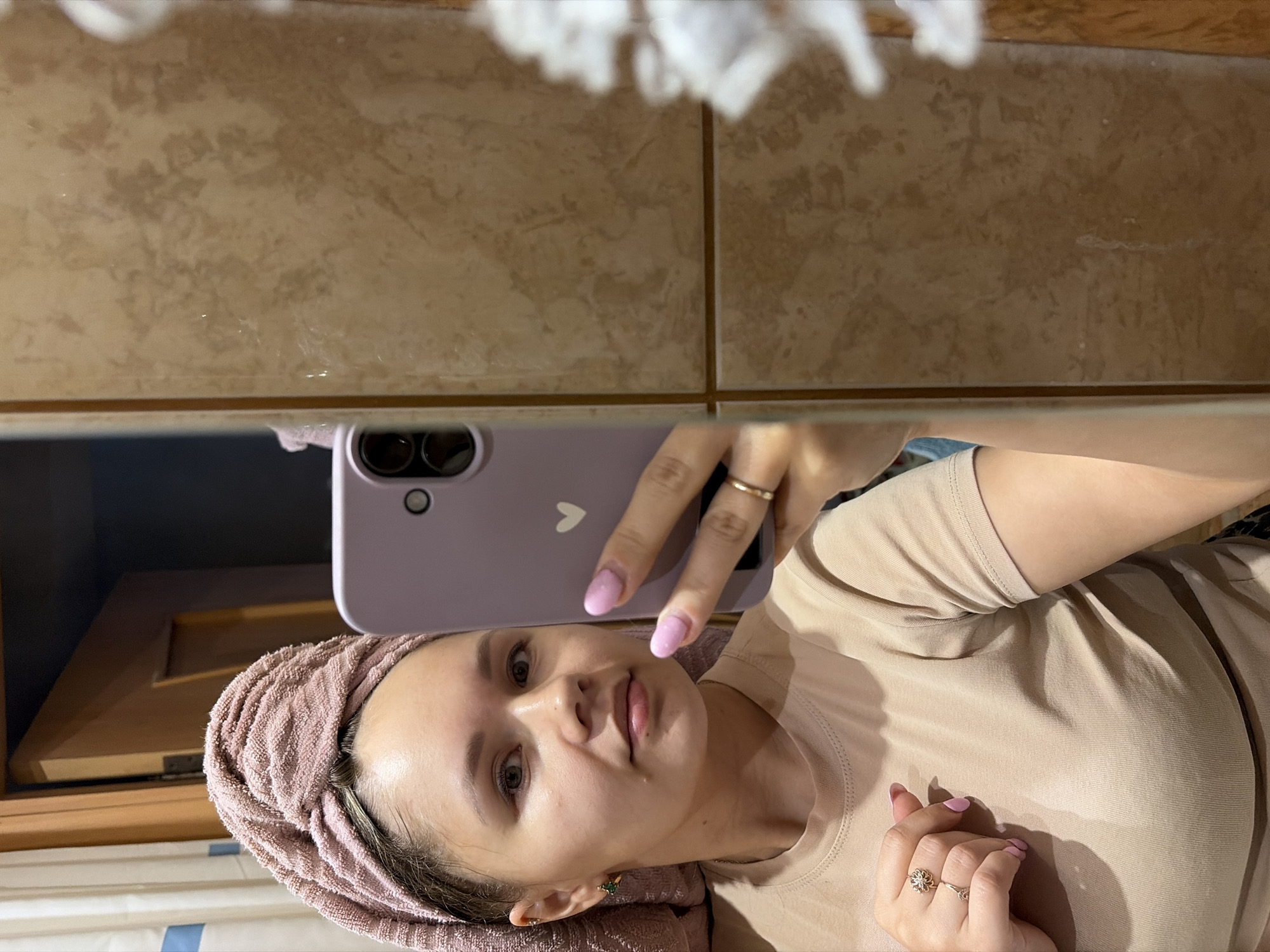 A woman is depicted taking a mirror selfie in what appears to be a bathroom, as indicated by the tiled wall. She is holding a light purple smartphone with a white heart sticker on its back and visible camera lenses, capturing her reflection. The image is oriented sideways, with the woman's head on the left and her body extending to the right.
The woman has light skin, dark hair tied back and covered by a light pink or dusty rose towel, suggesting she may have recently washed her hair or face. She is wearing a plain, light beige or tan short-sleeved t-shirt. Her face is in focus, showing a neutral expression, possibly a slight pout. She has light pink nail polish on her oval-shaped nails and wears two gold-toned rings on her right hand: a simple band on her index finger and a more ornate, flower-shaped ring on her ring finger.
The background visible in the reflection primarily consists of light brown or beige tiles with darker marbling, indicating a wall surface. Part of a wooden cabinet or furniture piece is also visible in the lower-left portion of the reflected image. There is no visible text in the image.