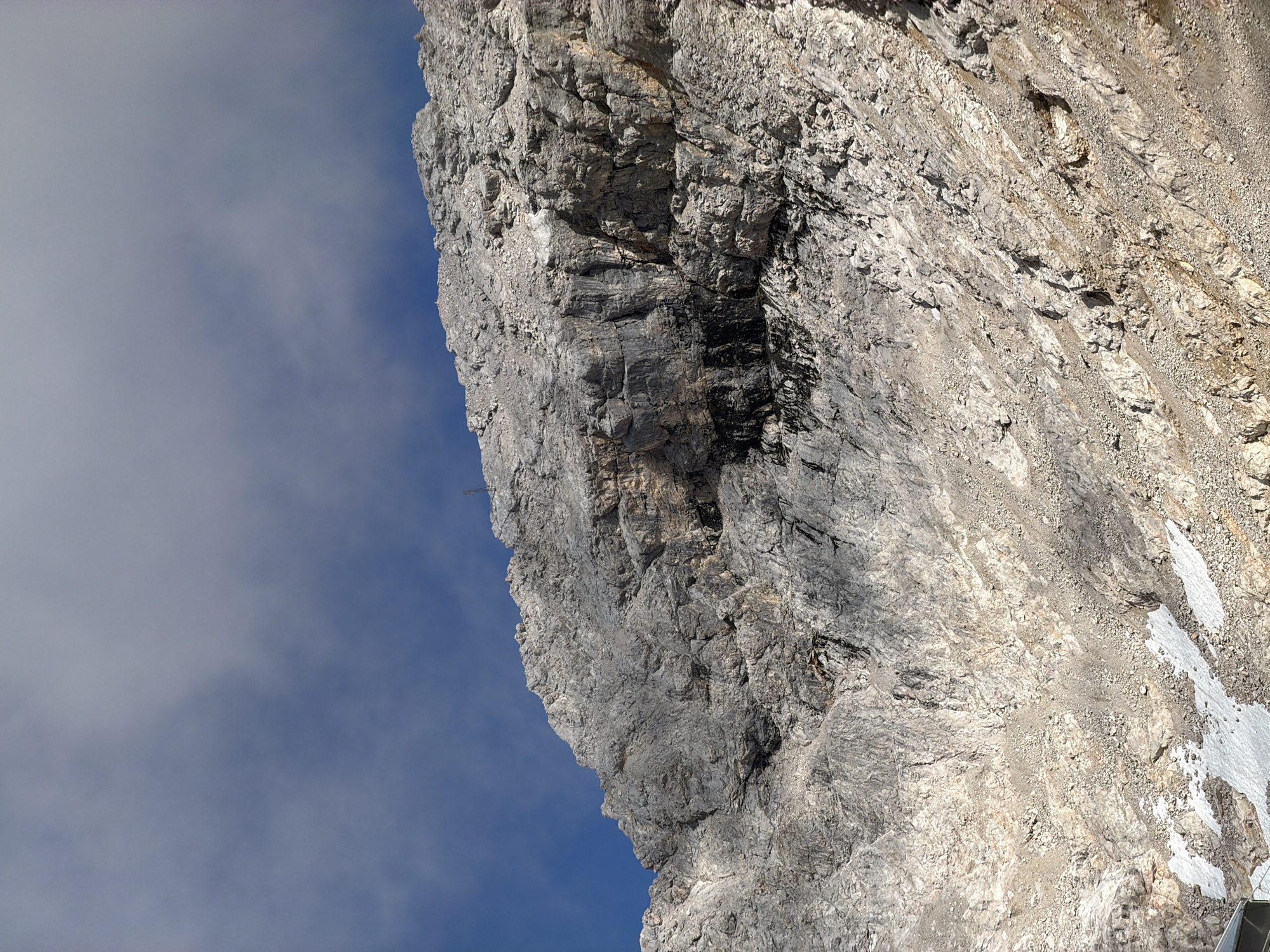This image captures a rocky mountain face under a sky with clouds. The terrain is steep and rugged, composed of gray and tan rock formations with scattered scree. Patches of snow are visible near the bottom of the image. The scene suggests a high-altitude mountainous environment. The location is the Zugspitze, the highest peak in Germany, located in the Wetterstein Mountains near Garmisch-Partenkirchen. There are no people or specific objects indicative of human activity, other than a faint structure at the bottom left edge, possibly part of a cable car station. The lighting suggests it is daytime, with sunlight casting shadows on the mountain.