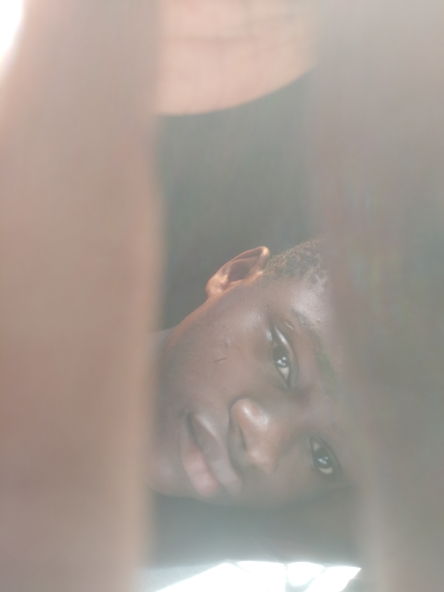 A close-up portrait captures a young individual, likely male, with dark skin, in Shangisha, Nigeria. The subject's face is prominently featured in the central, clearer part of the image, while the top and bottom are significantly blurred and overexposed with light brown and beige tones, possibly indicating out-of-focus hands or a very shallow depth of field. The person's head is tilted, and they gaze directly into the camera with a neutral, calm expression. Bright, natural light illuminates their face, creating highlights on their skin and a noticeable catchlight in their dark eyes. Their ear and short, dark hair are visible on the left side of the frame. The overall composition suggests an intimate or candid moment, possibly a selfie, taken during the daytime. No text is visible in the image.