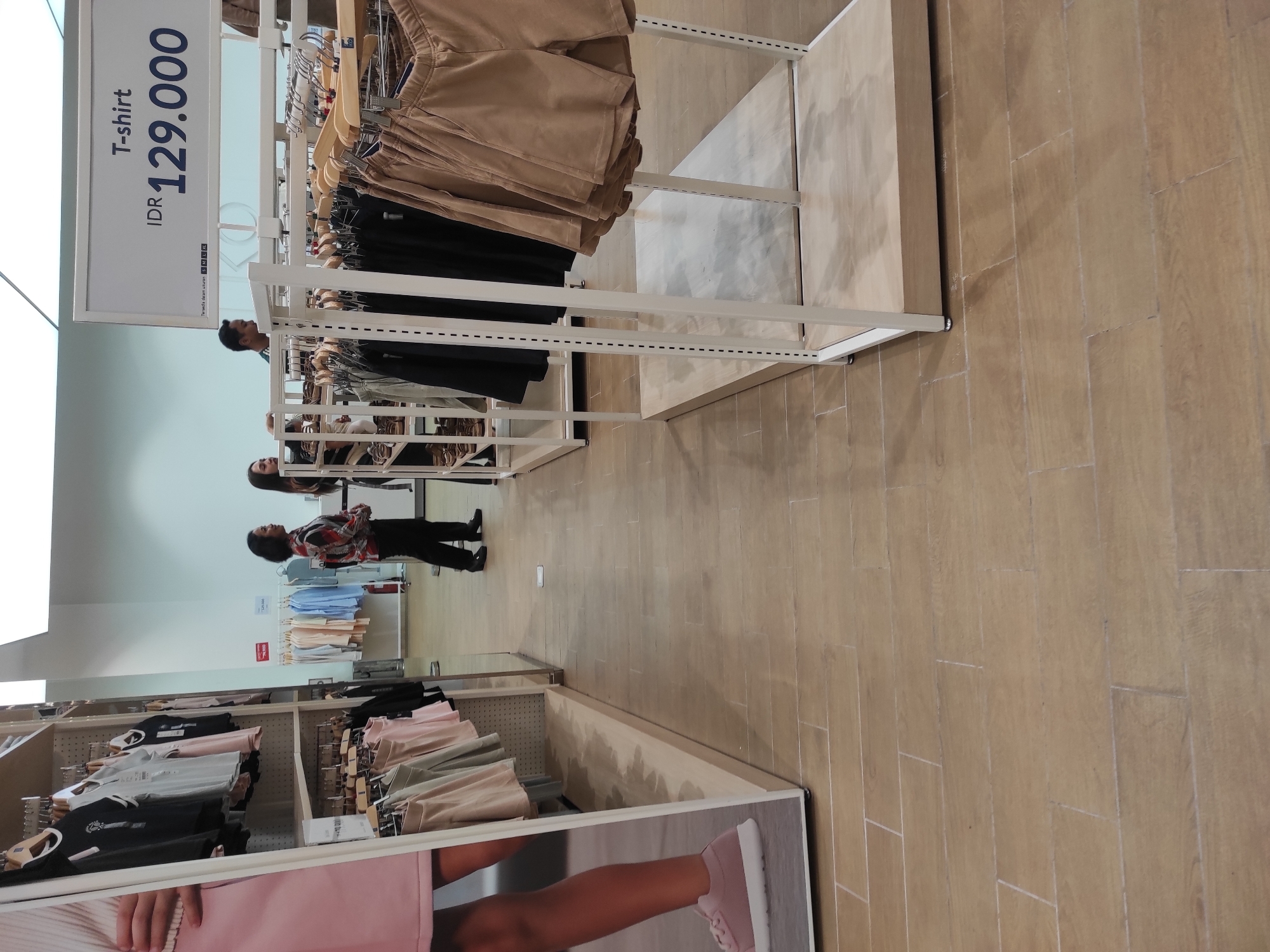 The image shows the interior of a clothing store, likely in Surabaya, Indonesia. In the foreground, a retail display features various clothing items, including t-shirts and shorts, on white shelving units. A large advertisement on the left showcases a person's legs wearing pink sneakers and a pink skirt. In the background, three individuals are visible. Two women stand near a rack of clothes, and a man stands behind them, looking towards the left. The store is illuminated by overhead lighting, and the flooring appears to be made of wood-look tiles. The overall environment is clean and organized, typical of a retail setting.