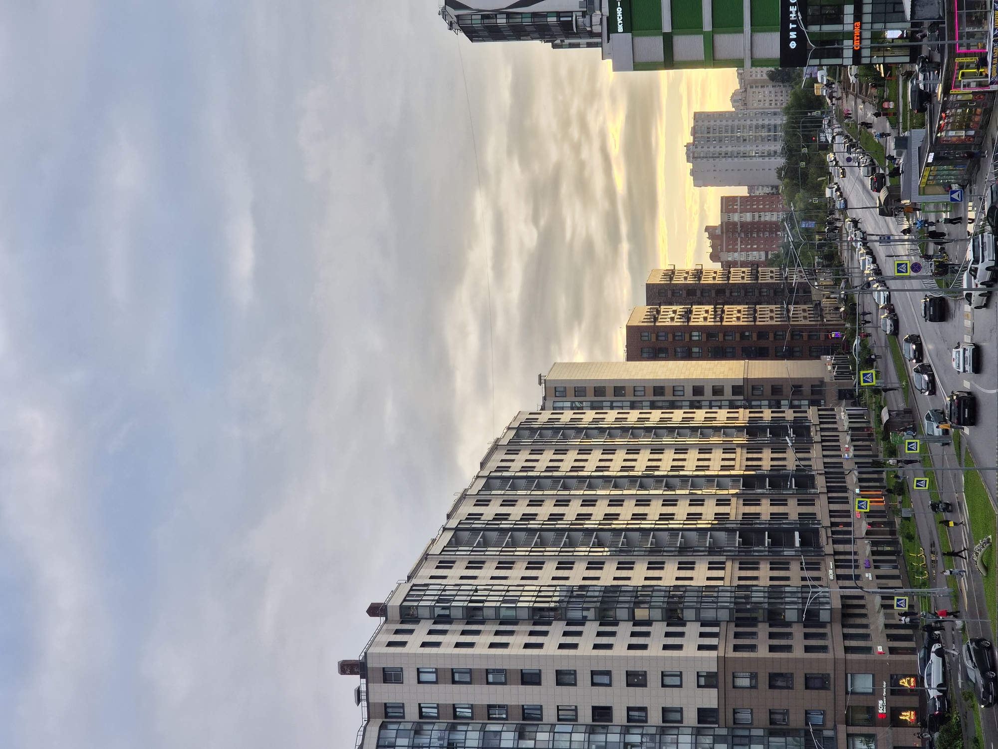 The image shows a wide street lined with multi-story apartment buildings and commercial establishments. On the left, a large, light-colored apartment building dominates the foreground, with numerous windows and balconies. Several cars, including a dark SUV and white sedans, are parked along the street and on the road. Pedestrians can be seen walking on the sidewalks and crossing the street, some with umbrellas, suggesting it might be raining or has recently rained, as the road appears wet. Yellow pedestrian crossing signs are visible.
Further down the street, more apartment buildings of varying architectural styles and colors are present. One building on the right has a prominent green facade with white text. A sign reading "ФИТНЕС" (Fitness) is visible on one of the buildings. The sky is overcast with warm tones of yellow and orange, indicating either sunrise or sunset. Traffic is moderate, with cars moving along the street. The scene is consistent with an urban environment in Kudrovo, Russia, a residential suburb of Saint Petersburg known for its modern apartment complexes and developing infrastructure.