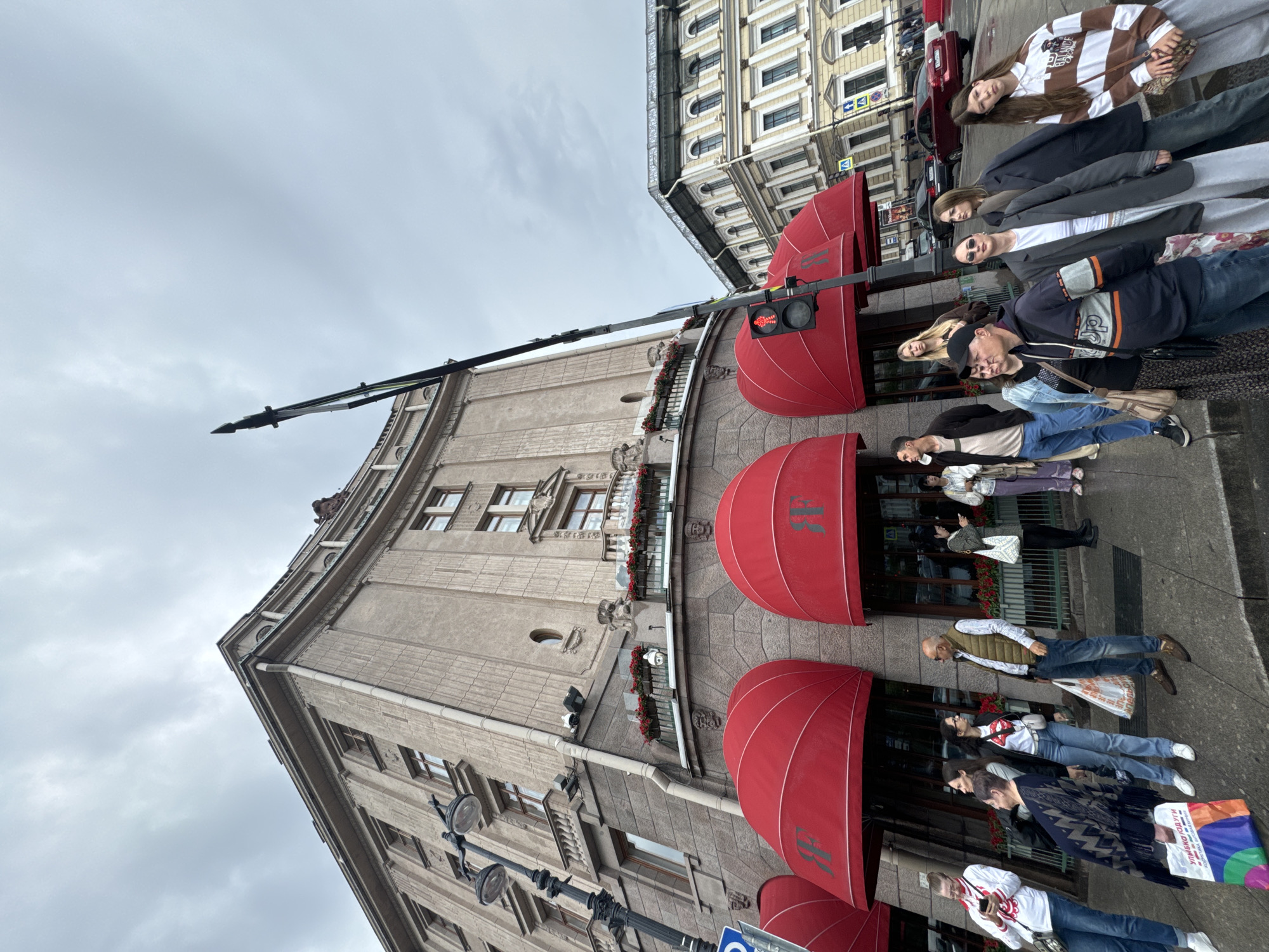 The image depicts a street scene in front of a building in Saint Petersburg, Russia, with a cloudy sky overhead. A group of people are standing on the sidewalk, some looking in different directions, suggesting they might be waiting or observing.
The building in the foreground features prominent red awnings above its entrance, with a stylized logo visible on them. The architecture is imposing, with multiple stories and balconies adorned with potted plants. A black lamppost with multiple lights stands to the left of the building.
Across the street, another building with a lighter facade and arched windows is visible. Traffic lights and street signs are present, indicating an urban environment. Several cars are parked or moving on the street.
The people in the image are dressed in casual clothing, and some are carrying bags. Their postures and gazes suggest a moment of pause or anticipation. The overall impression is one of a typical day in a bustling city, with the architecture and overcast sky adding a distinct sense of place.