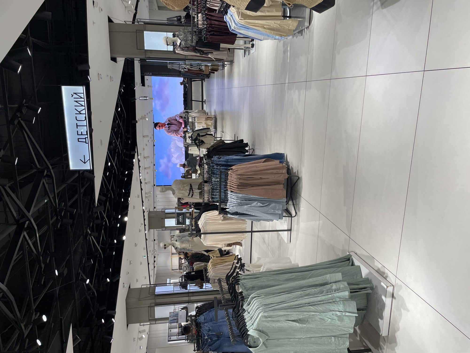 Here is a forensic description of the image:
**Location:** The image was taken in a clothing store in the city of Саларьево, Russia.
**Setting:** The scene is an aisle of the store, which is filled with racks of clothing. The floor is white tile. Above, the ceiling is a network of black architectural elements and spotlights. There is a sign, written in Russian, with an arrow that points left, saying "Детский" (which translates to "Children's"). In the background there is a large display screen, showing an image of a man against a cloudy blue and white background.
**Objects:**
* Racks of clothing in a variety of styles and colors
* A sign with an arrow
* White tile floor
* Large display screen
* Black structural supports
**Inferences:**
Based on the clothing and the sign, it is likely that this is the children's section of the store.