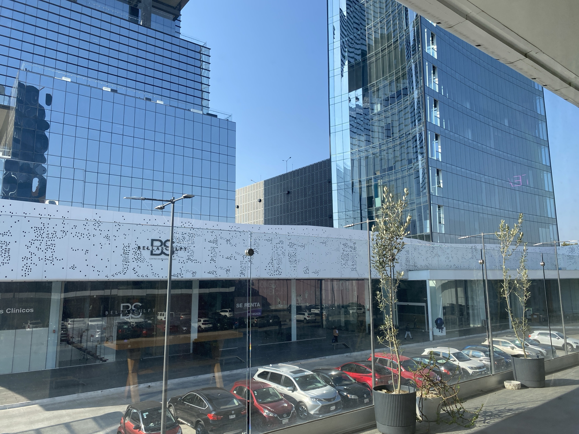 The image shows a view from inside a building overlooking an urban scene in Puebla, Mexico. Two modern, high-rise buildings with reflective glass facades dominate the skyline on the left and right. A lower, rectangular building sits between them, featuring a white facade with a pattern of perforations and the words "BELLASCULPT CENTRO DE BELLEZA Y SALUD". Below this building, storefronts are visible behind large glass windows, with cars parked along the sidewalk in front. Potted trees line the foreground, partially obscuring the view. The reflection of the camera-person is visible in the glass window.