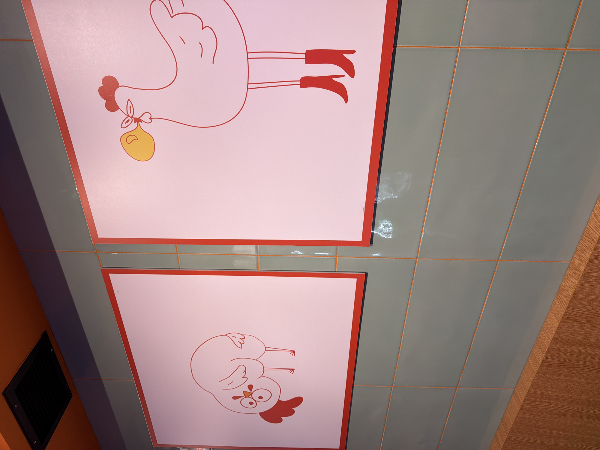 The image shows a section of a building interior. Two square panels with cartoon drawings of chickens are visible on a ceiling, placed among gray tiled sections. One panel features an upright chicken carrying a yellow sack with its beak, wearing red shoes. The other panel shows a chicken with an angry expression. The ceiling is made of rectangular gray tiles. Adjacent to the tiled ceiling is a wood-patterned surface. On the left side, there is a portion of an orange-painted wall with a black vent. The location is likely a restaurant or a similar establishment based on the decor and is found in Sofia, Bulgaria.