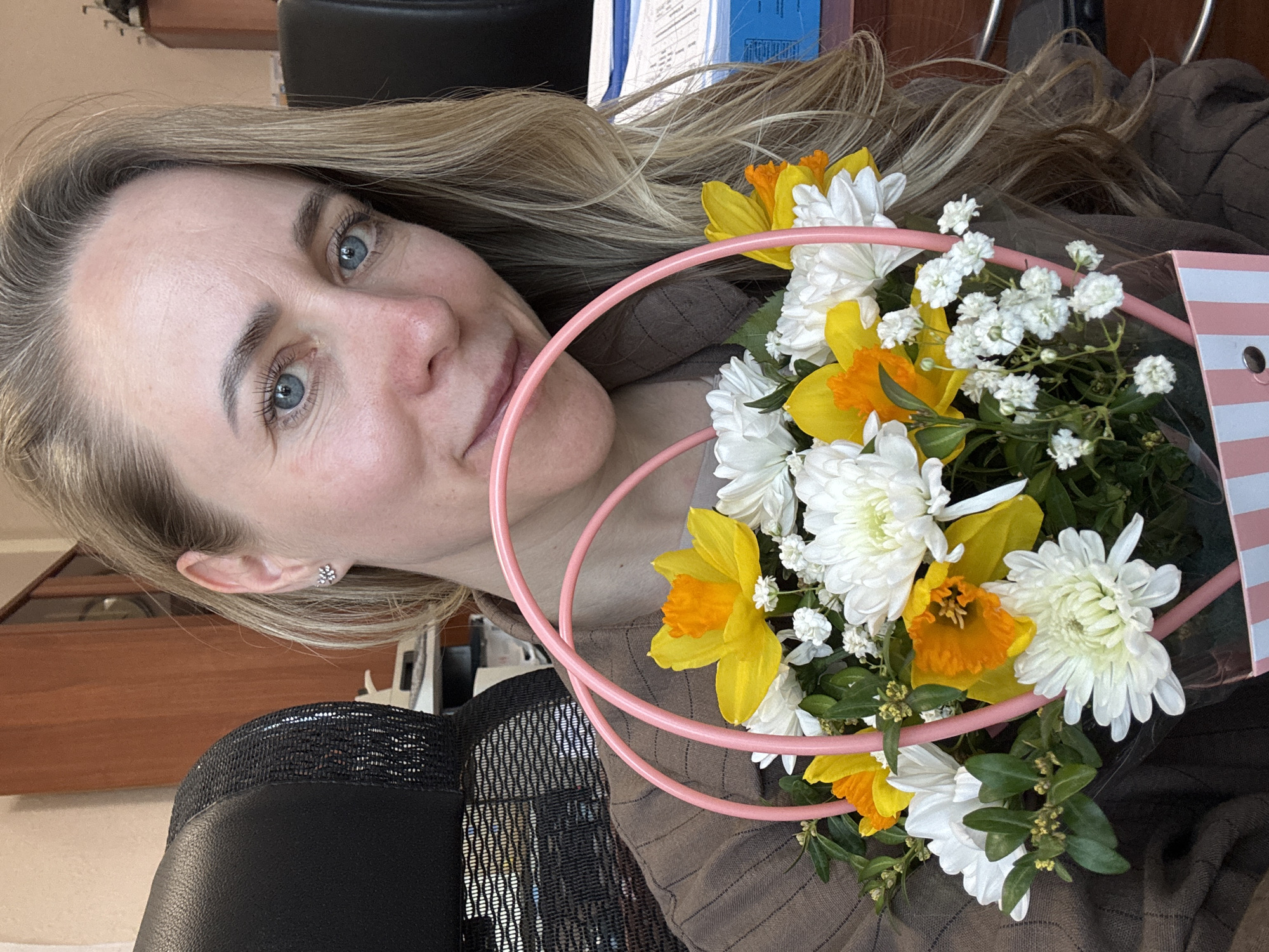 A woman with fair skin, blonde hair, and blue eyes is holding a bouquet of flowers. The bouquet contains white chrysanthemums, yellow daffodils, and white baby's breath, arranged in a round, pink frame. The woman is wearing a brown striped shirt. She is sitting in a black office chair, visible in the lower left of the frame. A wooden desk and some paperwork are partially visible in the background. It is not possible to determine the exact location in the city of Chaikovsky, Russia, from the image.