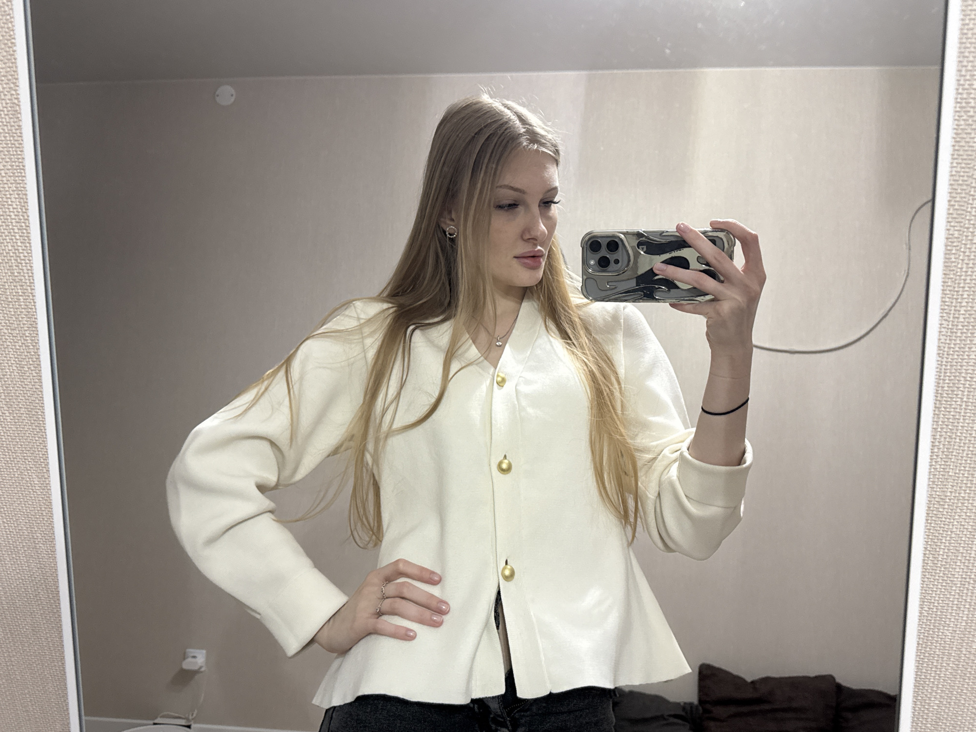 A young woman is taking a selfie in a mirror. She has long blonde hair and is wearing a white jacket with gold buttons and dark jeans. She is holding a smartphone with a camouflage case. The background shows a plain wall and a glimpse of furniture. The location is likely Perm, Russia.