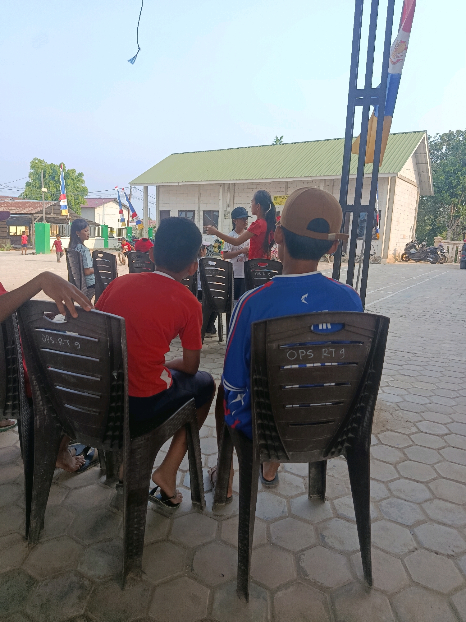 In Swarga Bara, Indonesia, an outdoor gathering is taking place in front of a building with a green roof and white brick walls. Several children and adults are present, some sitting on plastic chairs, others standing and interacting. One young girl in a red shirt appears to be addressing the group, while a boy wearing a tan baseball cap sits facing away from the camera. The ground is paved with hexagonal tiles. Flags are visible, and a motorcycle is parked in the background. The scene suggests a community event or educational activity.
