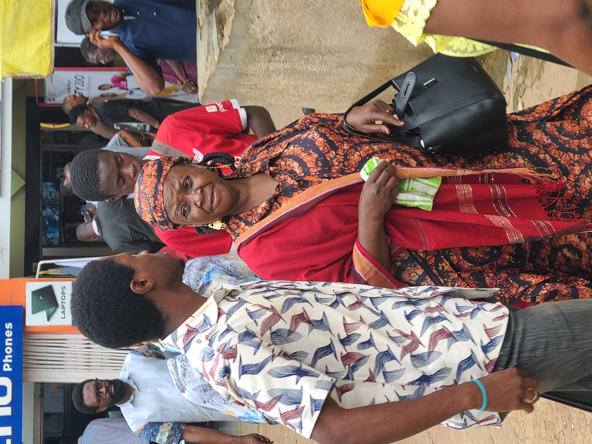 In Jos, Nigeria, a street scene unfolds with a medium shot of people in motion. A man in a patterned short-sleeved shirt, with a blue bracelet and a ring on his finger, stands in profile, looking to his left. To his right, a woman is captured from the waist up, wearing a headscarf, a red shawl over a black and orange patterned dress, and large gold earrings. She holds a black handbag in her right hand and a green and white striped cloth in her left. Behind them, other individuals are visible, including a man in a red shirt and a man with glasses and a beard in a light-colored shirt. The background suggests an outdoor marketplace or commercial area, with storefronts and signage, including a prominent "Phones" sign. The ground is rough and dusty, indicative of a typical Nigerian urban environment.