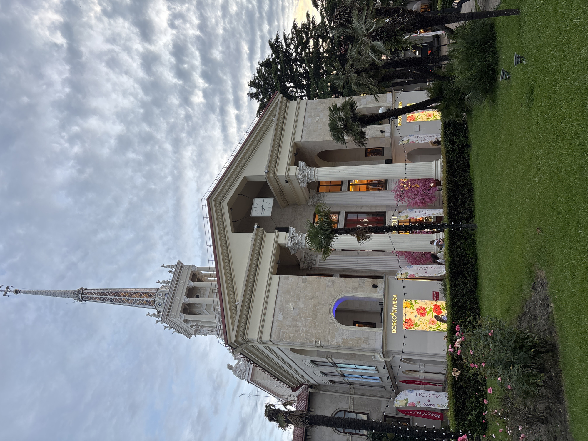 The image shows the Sochi Sea Port, a neoclassical building in Sochi, Russia. The port features a tall, slender spire with a clock face and decorative elements near its base. The building itself has a grand facade with columns and arched windows. In front of the building, there is a well-maintained green lawn with rose bushes and palm trees. Several vertical banners are displayed, some with floral designs and the "BOSCO RIVIERA" logo. A few people are visible, including a couple standing near a shop window and a person standing by a display of pink flowering branches. The sky is overcast with dramatic clouds, suggesting it might be dusk or dawn. Lights are visible in the shop windows, and strings of lights are draped along the facade.