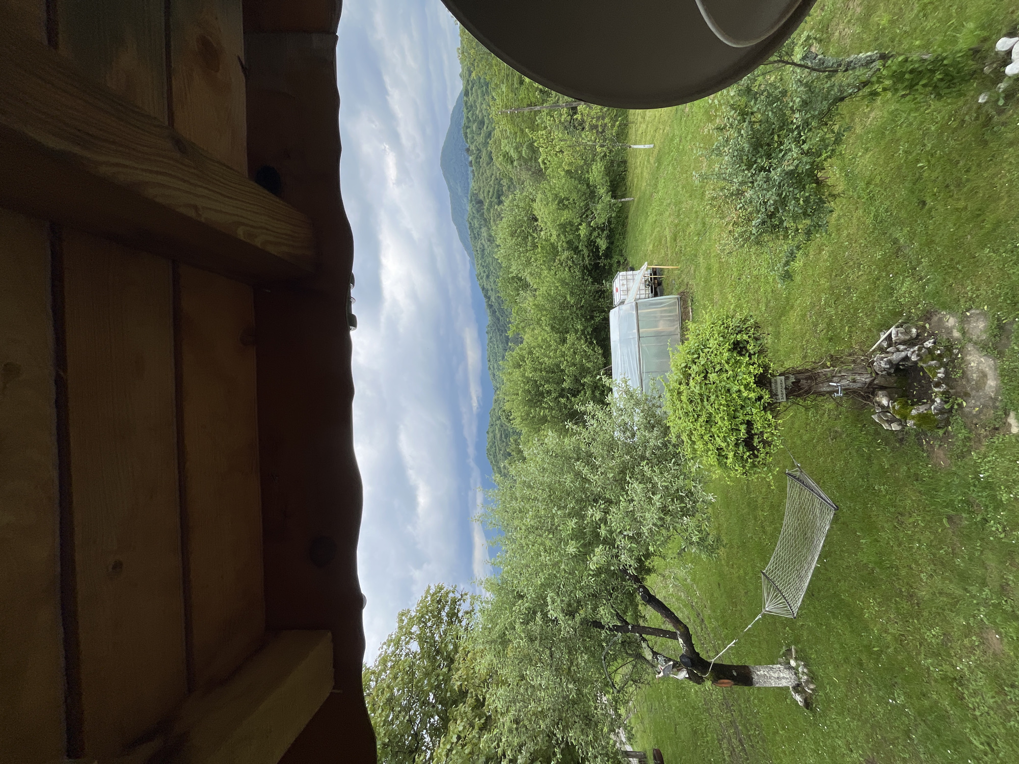 From within a wooden structure, a landscape is visible. A mountain range under a cloudy sky forms the backdrop. In the foreground, a green lawn hosts a greenhouse and a hammock suspended between trees. The setting is Shopite, Bulgaria.