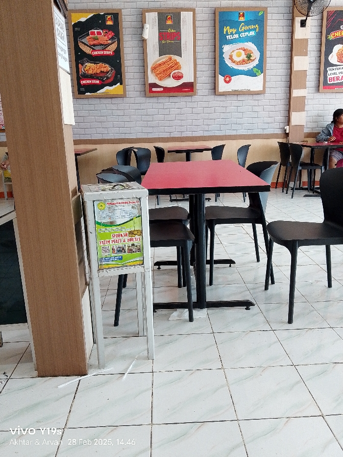 The scene is an indoor dining area, likely a restaurant or cafe. The location is identified as Patrol, Indonesia, at coordinates -6.318 latitude, 107.986 longitude. There is a red table with a red tabletop and four black chairs, two of which are empty. A white donation box, advertising “Sumbangan Yatim Piatu & Dhuafa,” is positioned to the left of the table. A wall with white brick pattern is visible behind the table, with posters promoting various menu items. The floor is tiled and there is a clear view of a portion of the floor in the foreground. One plastic straw lies on the floor. The interior appears to be clean and well lit. A second table with three chairs is visible in the background. The table is empty, and there is no one in the background other than the person holding the camera. The photo was taken on a smartphone by someone standing to the left of the table. The image was taken on February 28, 2023 at 14:46. The photo was taken using a Vivo Y19s.