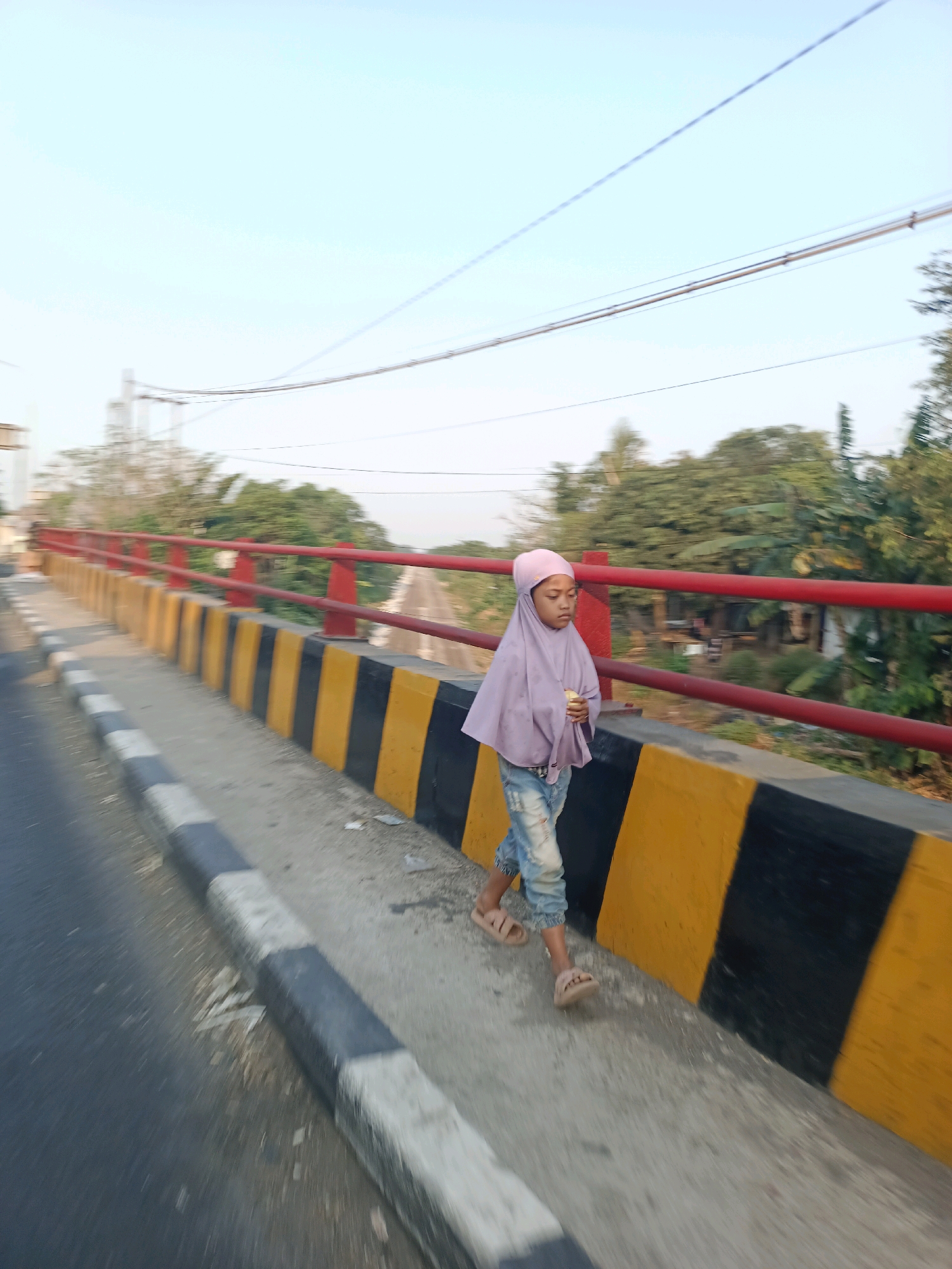 A young girl wearing a purple hijab and distressed denim jeans walks along the sidewalk of a bridge in Karawang, Indonesia. She is holding a yellow object, possibly food, in her right hand. The bridge has a red railing and a barrier with alternating yellow and black stripes. The road is to her left, and the background shows trees and buildings. The sky is clear and light blue.