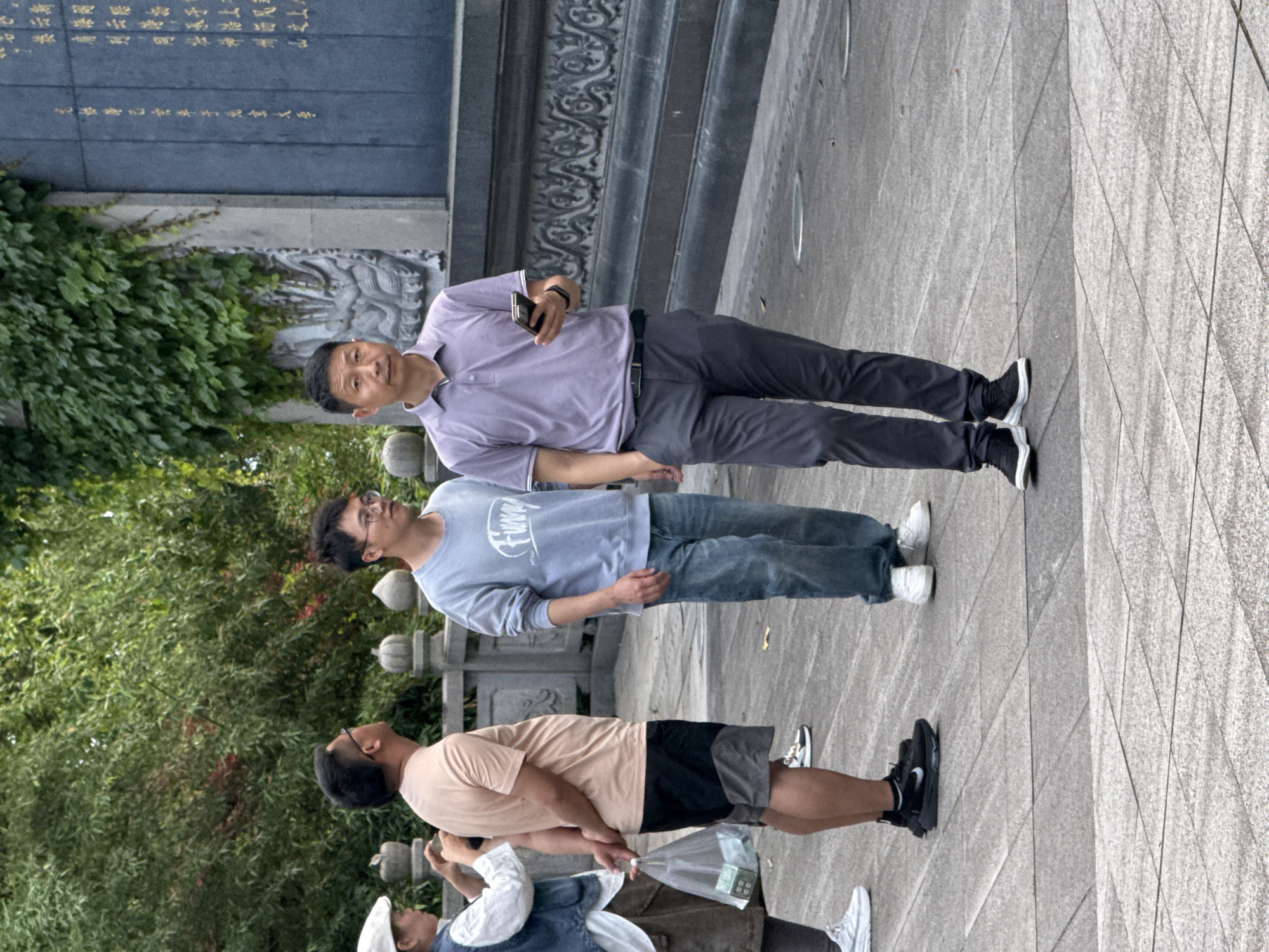 The image shows three people standing on a grey tiled walkway next to a low, ornate stone fence bordering a lush green area. In the background, a tall stone structure is visible, possibly a monument, with Chinese writing at the top and detailed carving beneath it.
The person closest to the camera is wearing a light brown t-shirt and black shorts with black athletic shoes. He is looking up. The next person is wearing glasses, a long-sleeve blue shirt with white lettering, and blue jeans. He is also looking up. The third person is wearing a purple polo shirt, grey slacks, and black shoes. He is holding a black phone. The image was most likely taken in the Liangjiang, China region, based on the architectural style and landscape.
