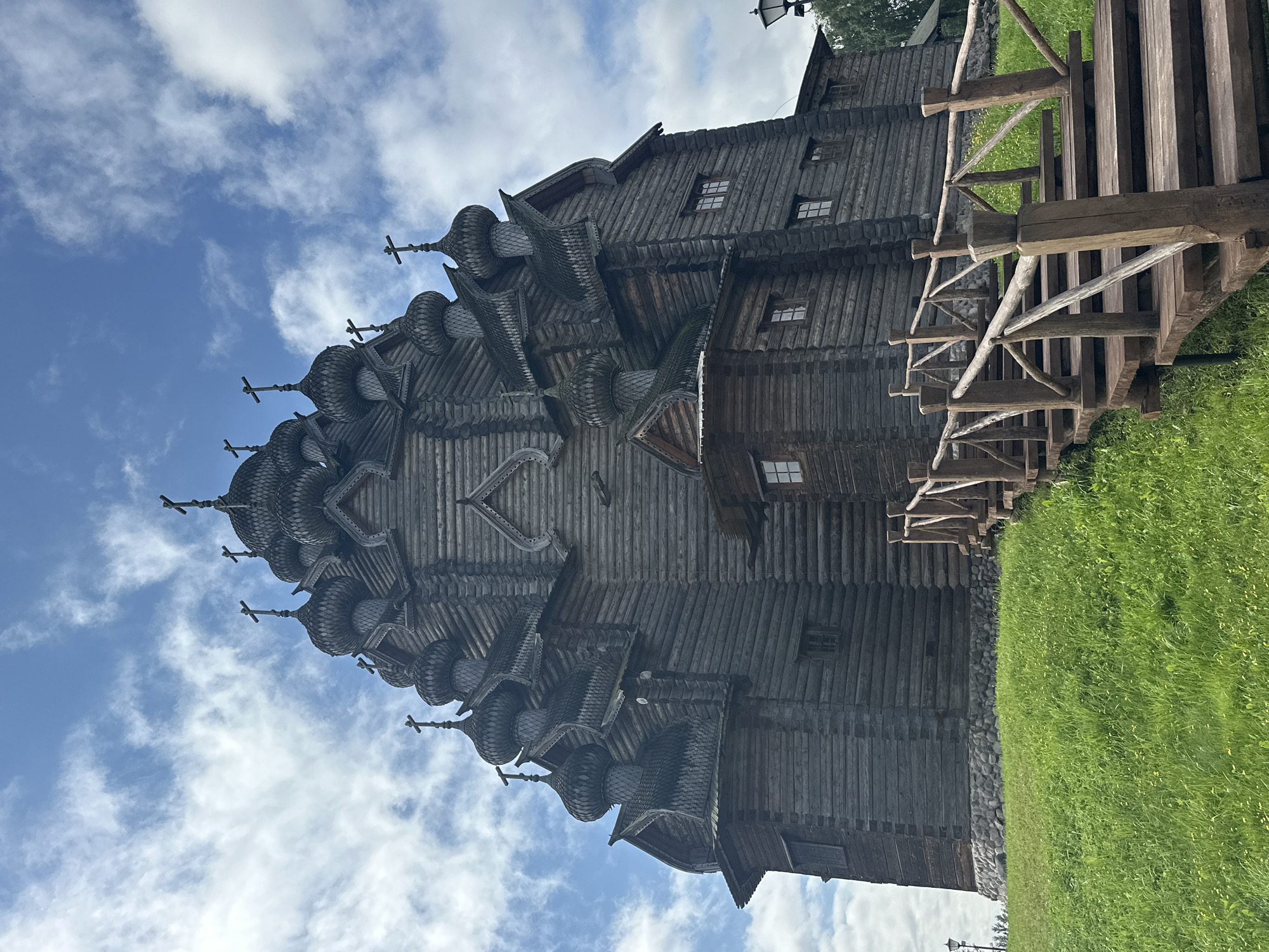 In Nevskii Parkleskhoz, Russia, a large, multi-domed wooden church stands tall against a bright blue sky with scattered white clouds. The church is constructed entirely of dark, weathered logs and features numerous onion-shaped domes, each topped with a cross. A wooden staircase with rustic railings leads up to the church from the foreground. The church is situated on a grassy hill, with some stone detailing at its base. The overall scene is serene and highlights traditional Russian wooden architecture.