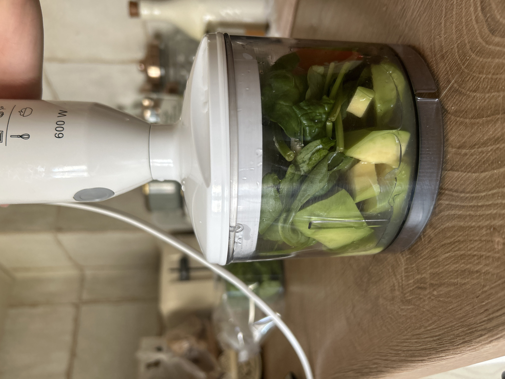 An immersion blender with a "600 W" label is positioned over a clear plastic chopping container. The container is filled with chopped avocado, spinach, and possibly a piece of tomato. The blender's white body and cord are visible. The container sits on a wooden countertop in what appears to be a kitchen. In the blurred background, there are kitchen tiles, jars, and a bottle. The scene does not provide any information about the city of Plovdiv, Bulgaria.