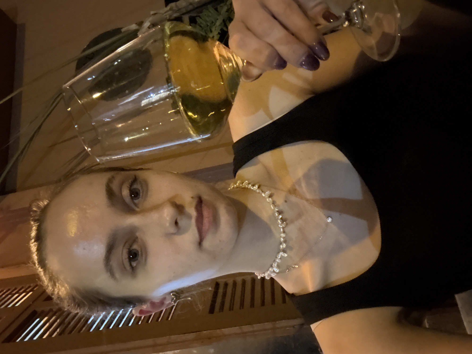 A woman is holding a wine glass filled with white wine. She is wearing a black top and a silver necklace. The environment appears to be a bar or restaurant, indicated by the wooden construction and plants in the background. The city of Tel-Aviv, Israel, is not indicated in the image.