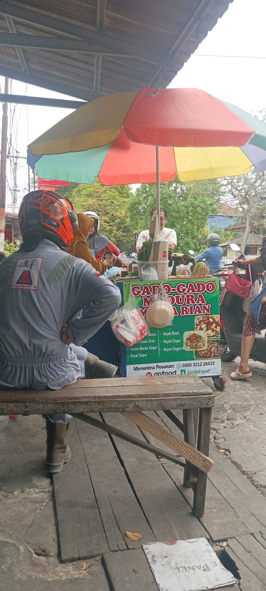 The image captures an outdoor scene in Balikpapan, Indonesia, likely a street food stall. In the foreground, a person wearing a grey uniform and an orange and black helmet is seated at a wooden table. The uniform has a logo indicating "PT. BALIKPAPAN READY MIX". Another person, wearing a hijab and a grey scarf, is seated to the left of the uniformed individual.
Behind them, a food cart is visible, covered by a multi-colored umbrella (red, yellow, green, and blue). The cart displays a sign advertising "GADO-GADO MADURA" and lists various menu items such as "Gado Telur", "Gado Gado Cingur", "Pecel Sayur", "Rujak Buah", and "Rujak Cingur". There are also images of the food items on the sign. A vendor, a man wearing glasses and a light-colored shirt, is attending the stall. He is seen interacting with customers and preparing food. Several plastic bags containing food items and containers are placed on the cart.
In the background, trees and some buildings are visible, suggesting an urban or semi-urban environment. A motorcycle is parked to the right of the food cart, with a person on it wearing a blue jacket. The ground is a mix of concrete and wooden planks, appearing weathered. The overall atmosphere suggests a casual, everyday setting where people are purchasing food.