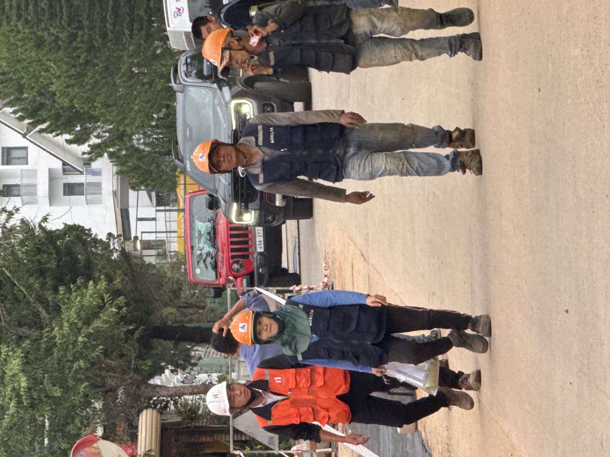 In Da Lat, Vietnam, a group of construction workers walks along a dirt road. The primary subjects are four men, all wearing hard hats and vests. The man on the far left wears a white hard hat and an orange safety vest over a black shirt, black pants, and brown boots. To his right, another man in a blue shirt and dark pants is partially obscured. The third man from the left wears an orange hard hat, a dark blue vest with "ADELTA" written on it, light blue jeans, and dark boots. He carries a clear plastic bag in his left hand. The man on the far right, also wearing an orange hard hat, has on a dark vest, light-colored jeans, and dark boots. He appears to be smoking. Behind the workers, several vehicles are visible: a red Jeep Wrangler with license plate "49A-377", a dark SUV, and a white delivery van. The background shows trees, a multi-story building with balconies, and residential structures. Construction barriers and tapes are present along the side of the road. The ground is uneven and dusty.