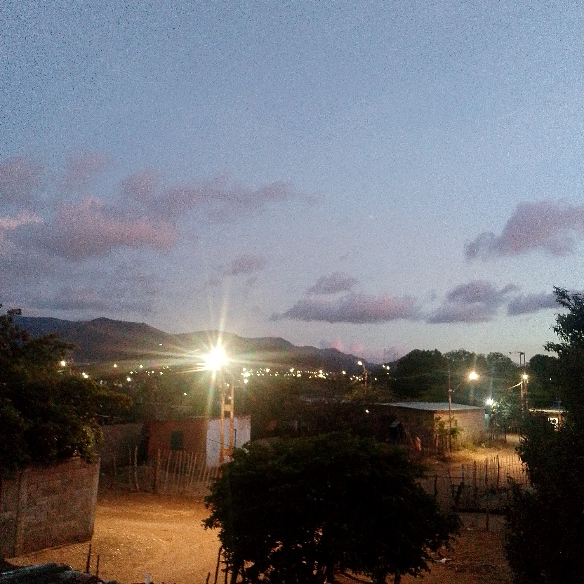 The image shows a street scene at dusk in Altagracia, Venezuela. The sky is a light blue-gray with some pink-tinged clouds. In the background, there are mountains. Street lights illuminate the scene, casting a bright glare. There are houses and trees visible on either side of the dirt road. The area appears residential. There are no people visible in the image.