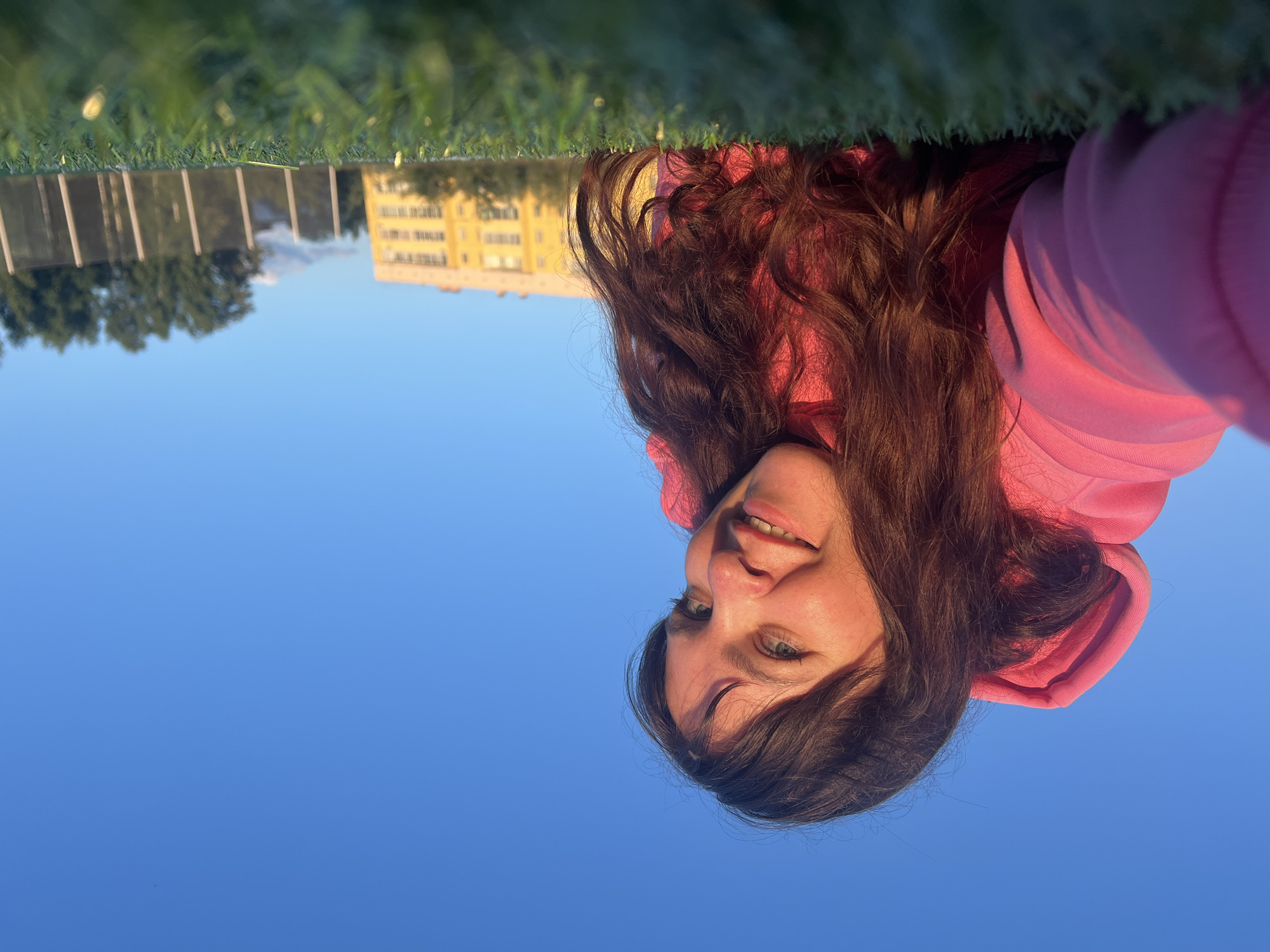 The image features a young woman with long, dark brown hair and blue eyes, wearing a pink hoodie. She is looking up at the camera with a smile. The background is a clear blue sky, and the reflection of grass and buildings is visible as if the image is taken from a mirror or a reflective surface. The buildings appear to be residential, and the presence of grass suggests an outdoor setting. The lighting indicates it might be daytime, possibly late afternoon or early evening, given the warm tones on the buildings and the grass. The woman's expression conveys a sense of happiness and playfulness. No text is visible in the image.