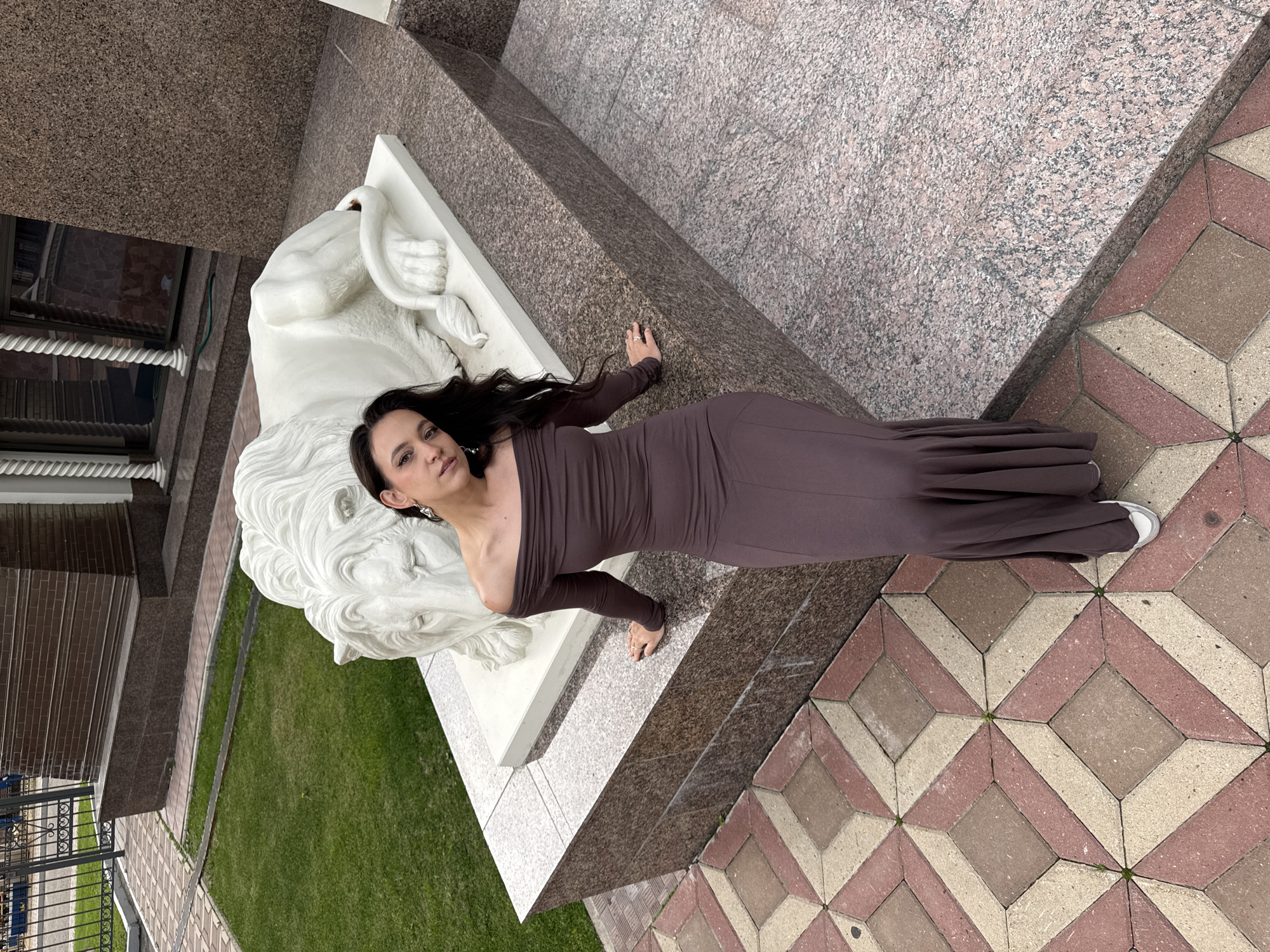 In Kazan, Russia, a young woman with long, dark hair and dark eyes is photographed outdoors. She is wearing an off-the-shoulder, long-sleeved, dark brown maxi dress and white sneakers. The woman is posing next to a white sculpture of a resting lion. She is positioned on a granite pedestal, leaning with her left hand on its surface. The pedestal is situated on an outdoor paved area with a geometric pattern of red and beige tiles. Behind the woman and the lion sculpture, a manicured green lawn is visible, bordered by a pathway and a decorative metal fence. Further in the background, a modern building with brick and stone facades can be seen. The lighting suggests it is daytime, possibly overcast.