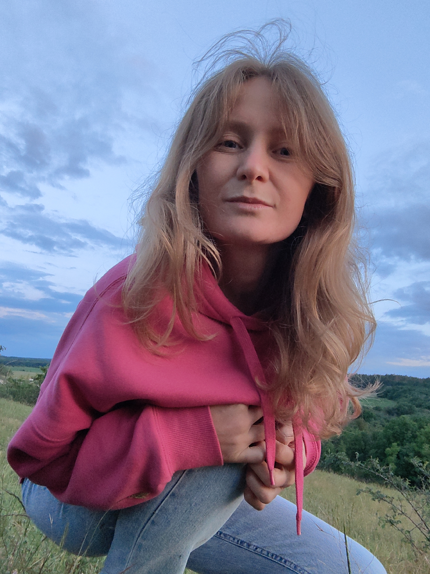 A woman with blonde hair, wearing a pink hooded sweatshirt and blue jeans, is crouching outdoors. She is looking towards the camera. Her hands are clasped in front of her. The background shows a landscape of rolling hills covered in green trees and dry grass, under a cloudy blue sky. The image appears to have been taken during the evening or early morning. The location is in the vicinity of Зуйское сельское поселение, Ukraine.