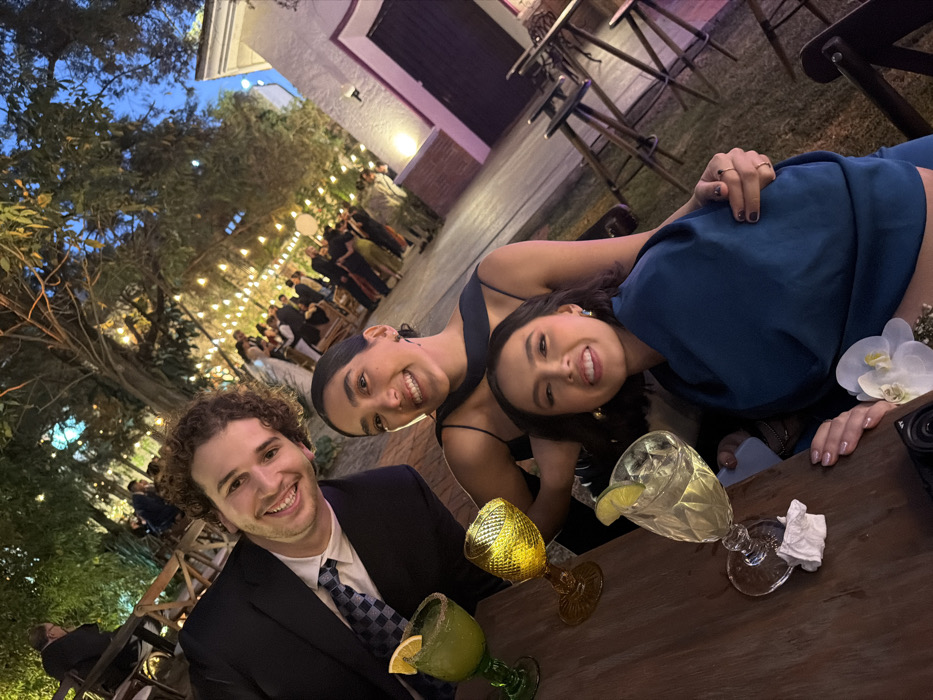 Three people pose for a photo at an outdoor event. A man in a suit and tie stands to the left, smiling. Two women are next to him; one wears a black dress with thin straps, and the other is in a blue outfit, both looking at the camera with smiles. A table in front of them holds three colorful, garnished drinks. String lights are visible in the background, along with trees and other people attending the event.