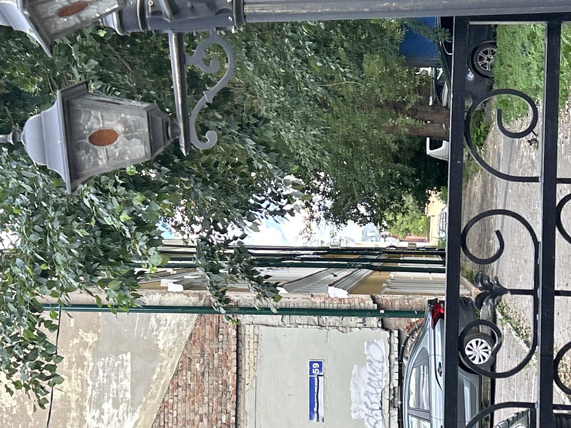 This image depicts a street scene in Perm, Russia, with a focus on architectural elements and vehicles. In the foreground, a black wrought-iron fence with decorative scrollwork runs horizontally across the frame. Behind the fence, a grey sedan is parked. To the left, a building with a weathered brick and stucco facade is visible. A street sign in blue and white indicates "Маузерская 59" (Mauser Street 59). A green downspout runs vertically along the building. In the middle ground, a row of trees with dense green foliage partially obscures the view of other buildings and a paved walkway or road. A vintage-style street lamp with a dark metal frame and amber-colored glass panels is prominent in the upper right quadrant of the image. The background reveals more trees and what appears to be the side of a blue structure, possibly a truck or building cover. The overall scene suggests an urban environment with older architecture and residential or mixed-use buildings.
