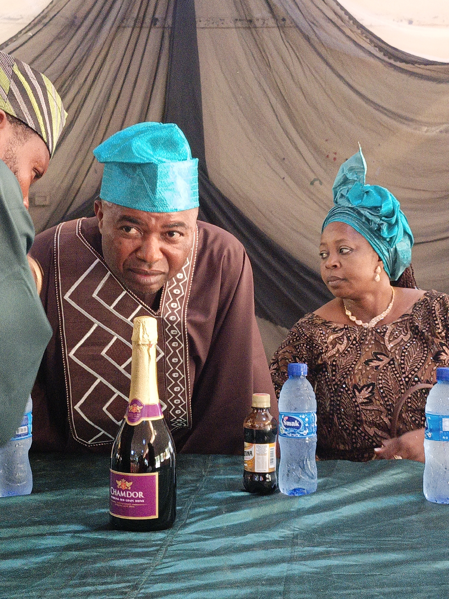 Fagge D2, Nigeria. A medium, eye-level shot depicts three individuals seated at a table covered with a teal-colored cloth. A man in a brown traditional outfit with geometric white embroidery and a turquoise cap sits in the center. A woman to his right wears a similar turquoise head wrap and a brown patterned dress, accessorized with a pearl necklace and earrings. To the left of the central figure, a portion of a man's face is visible, wearing a green and striped traditional cap.
On the table, a bottle of Chamdor red grape drink stands prominently in front of the man in the center. Several water bottles and a small brown bottle are also present. The background features draped black netting.