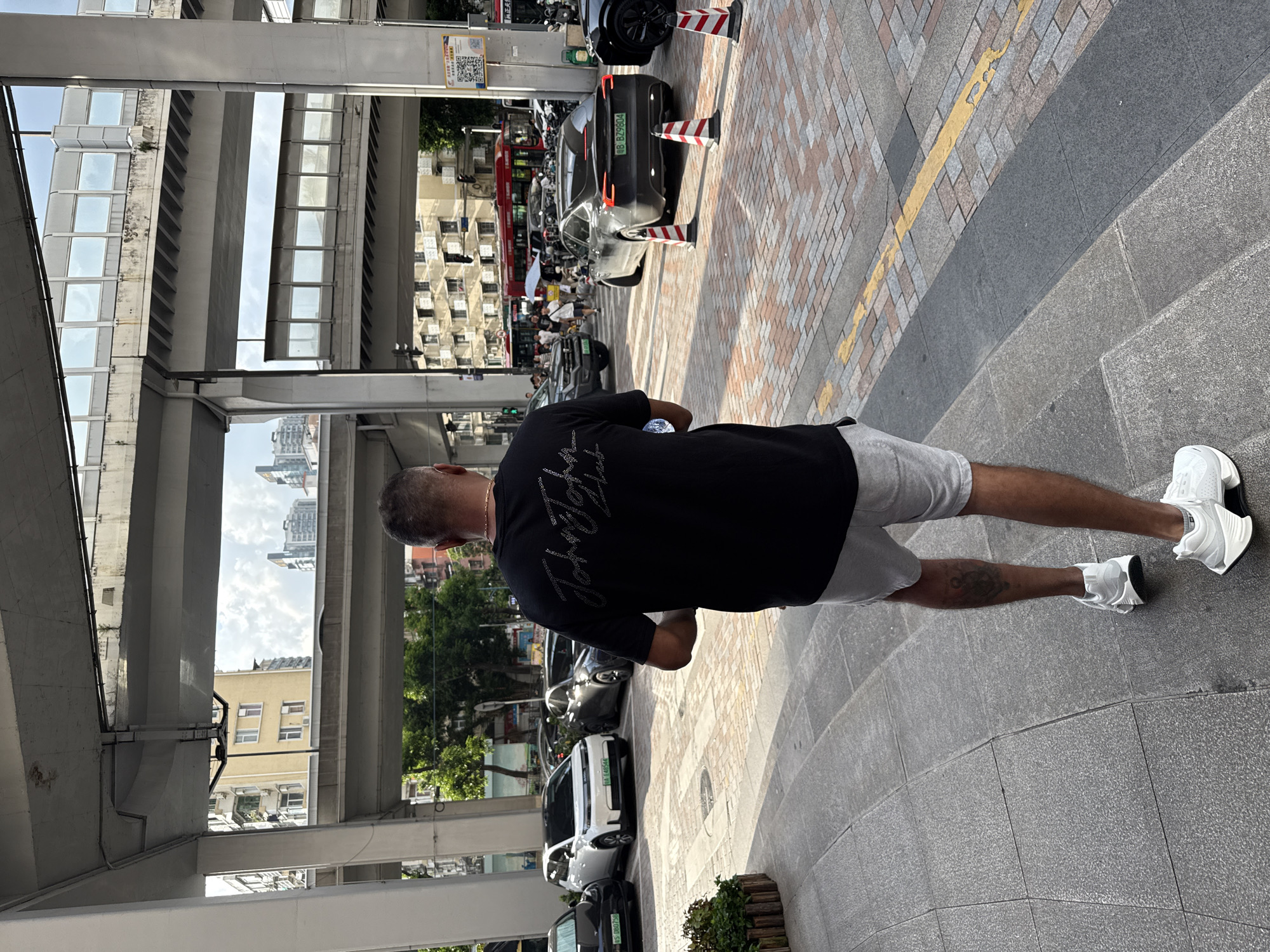A man is walking away from the camera on a sidewalk in Haizhu District, China. He is wearing a black t-shirt with a sparkly graphic on the back, grey shorts, and white sneakers. The man has a tattoo on his left calf. In the background, several cars are parked along the street, including a white electric vehicle and a black Tesla. An elevated highway passes overhead. The buildings in the background appear to be a mix of modern and older architecture. The street is paved with brick-like tiles.