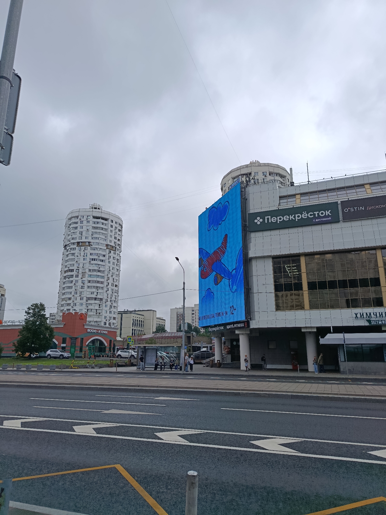 The image shows a street view in Moscow, Russia, on a cloudy day. In the foreground, there is a multi-lane asphalt road with white directional arrows painted on it. A yellow dashed line separates lanes on the left side of the road. In the mid-ground, there is a bus stop with several people waiting. Adjacent to the bus stop is a building with a large blue digital billboard displaying a cartoon airplane and clouds. The billboard also features text that includes "Перекрёсток" and "spirit.FITNESS", indicating retail establishments or services. To the left of the billboard, there is a round, white apartment building with many windows. Further to the left, a red building with the text "ВКУСНО – И ТОЧКА" is visible, which is a fast-food chain. In the background, other buildings and a street lamp are present. The overall scene depicts an urban environment with a mix of residential and commercial structures.