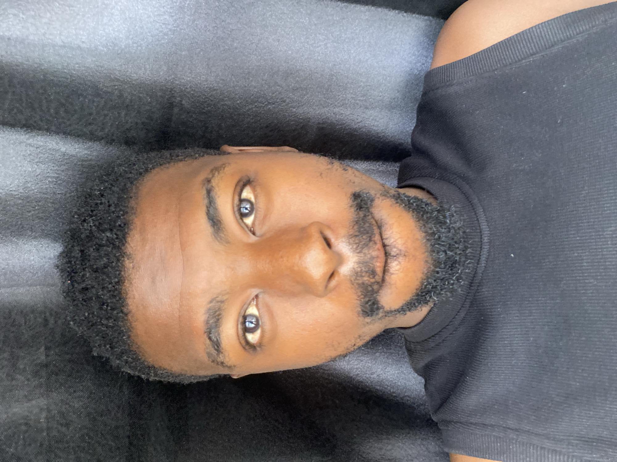 An adult man is captured in a close-up portrait, oriented horizontally as if lying down or reclined, looking directly into the camera. He has dark skin, short, dark curly hair visible around the edges of his face, and a full, well-groomed dark beard and mustache. His dark brown eyes are open and gaze steadily, reflecting a light source. He wears a plain black, ribbed t-shirt, the texture visible on the fabric around his neck and shoulders. The background is a uniform dark color, appearing to be a textured fabric or upholstery, possibly a car seat or a couch, with subtle folds. The lighting illuminates his face directly, highlighting his features. No specific activity beyond posing for the photo is discernible. There is no visible text. The location context for this image is Barakin Akawo, Nigeria.