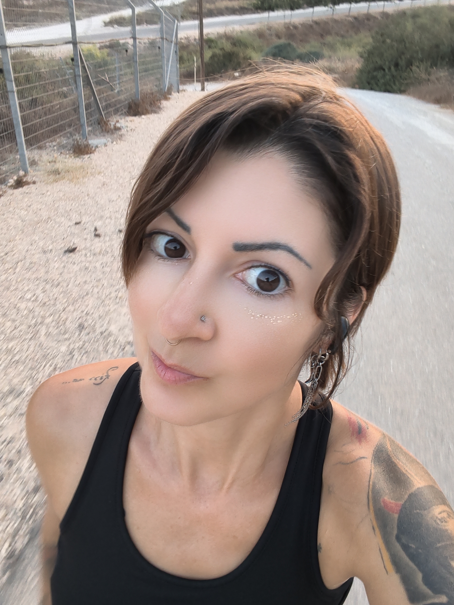 In an outdoor, arid environment, a woman with short, brown hair is captured in a close-up, selfie-style photograph. She is looking directly at the camera with wide, brown eyes, and her mouth is slightly open. Her skin is fair, and she is wearing makeup, including what appears to be glitter or shimmer on her cheekbones. She has a nose ring and a hoop earring in her left ear. She is wearing a black tank top, and tattoos are visible on her left shoulder and upper arm, as well as a small tattoo on her left collarbone. To her left, a metal fence with a concrete base runs along a dirt path. In the background, there is more arid vegetation and a paved road winding through a hilly landscape. The scene is consistent with locations in the Lachish Regional Council, Israel, particularly near border areas or agricultural land.