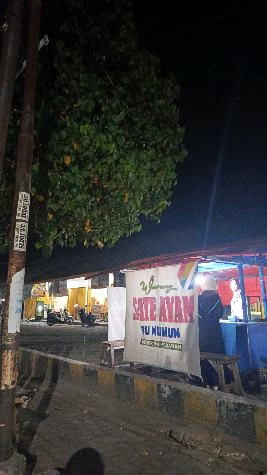 The image depicts a street scene at night in Glonggong, Indonesia. In the foreground, a makeshift food stall is set up, featuring a banner that reads "Warung Sate Ayam Bu Mumun" and "Menerima Pesanan" (Accepting Orders). Two individuals are visible at the stall, one wearing a helmet and facing away from the camera, and another, a woman, partially visible behind the counter. The stall itself is adorned with red and blue fabric. To the left of the stall, a utility pole with stickers is present, and beyond that, a row of shops with illuminated storefronts and parked motorcycles can be seen. The street is paved, and a concrete barrier with yellow and black markings separates the sidewalk from the road. Lush green foliage from a nearby tree overhangs the scene. The overall impression is of a bustling local eatery operating into the evening.
