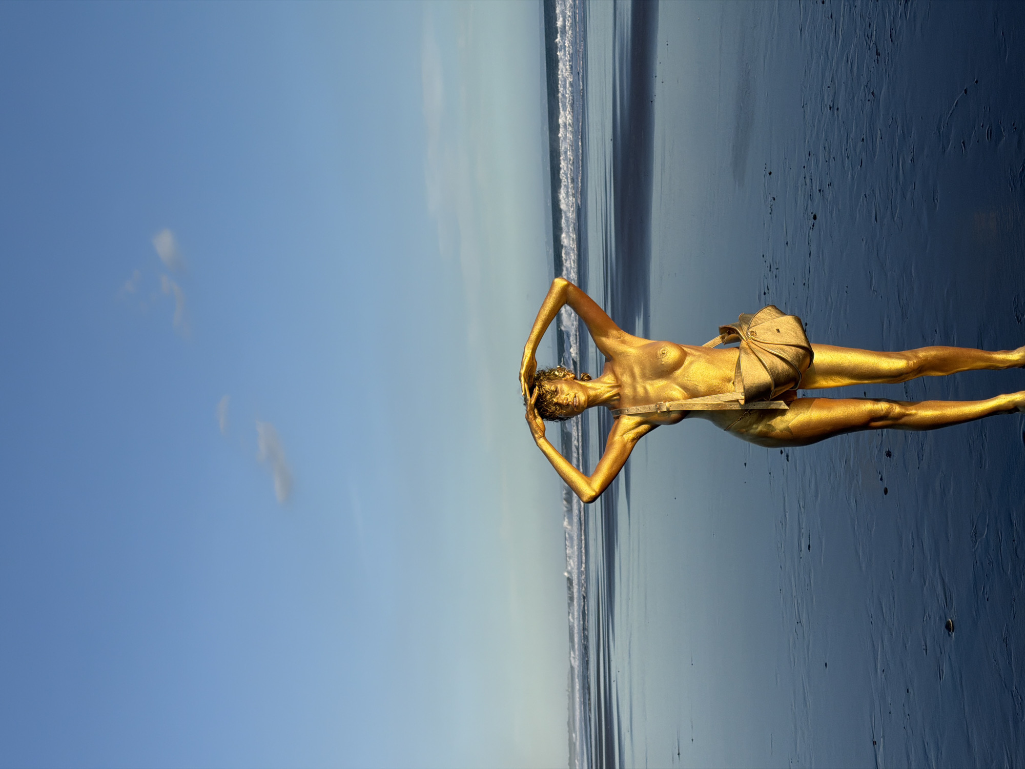 A golden-painted woman lies on a beach, facing to the right, with her hands resting on her head. The sky is blue with a few clouds. The location appears to be a beach in Kerambitan, Indonesia. The surface she lies on appears to be wet black sand, reflecting the sky. Ocean waves are visible in the background.