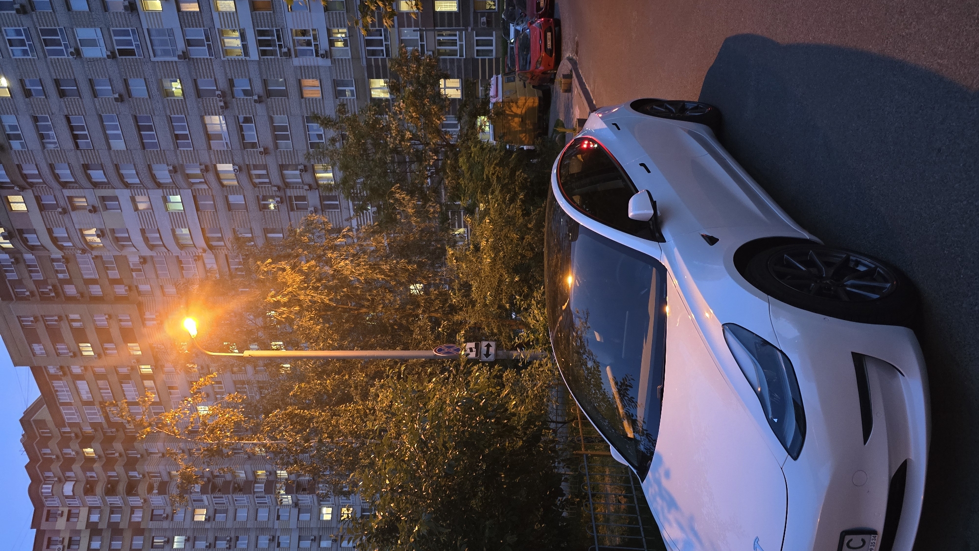 The image captures an urban residential scene during dusk or night, with a white Tesla sedan prominently parked on an asphalt street in the foreground. The Tesla features dark-colored wheels, and its pristine white body reflects the warm, orange light from a nearby streetlamp.
To the left, a towering multi-story apartment building stands, its numerous windows forming a complex grid. Many windows glow with internal lights, suggesting active residences. The building's architecture, with its repetitive window structures and external air conditioning units, suggests a modern urban environment.
Between the street and the apartment building, a line of dense trees and foliage is brightly illuminated by a powerful, warm orange streetlamp positioned in the mid-ground. The light casts a golden hue over the leaves. Further down the street, a red car, possibly a Ford Fiesta, and another vehicle are parked.
A traffic sign, featuring a red and blue "No Stopping/Parking" symbol atop a rectangular panel with vertical arrows indicating the restriction's scope, is affixed to the streetlamp pole. No people are visible in the tranquil, evening setting. The overall atmosphere is quiet and still. A partially visible license plate on the front of the Tesla shows the letter "C" followed by some numbers. This scene is consistent with an urban area in Kyiv, Ukraine.