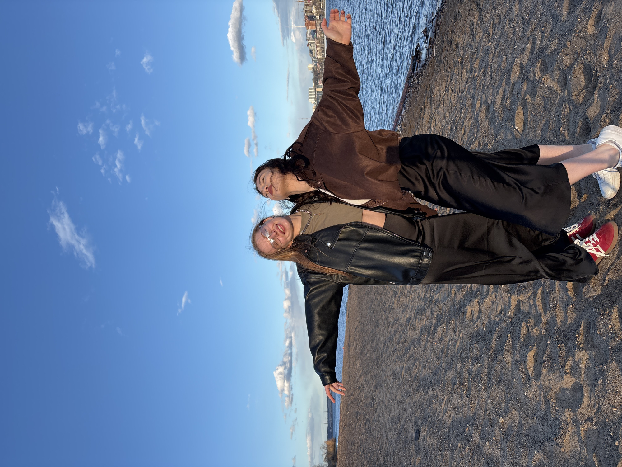 Two young women are captured in a horizontal, brightly lit photograph, posing on a sandy and gravelly shoreline next to a body of water. The image, if rotated correctly, would show them standing upright.
The woman on the left (when viewed as correctly oriented) has long, light brown hair and is wearing clear-framed glasses. She sports a black leather jacket over a green or khaki top, black wide-leg trousers, and red and white sneakers. A silver chain necklace is visible. She is smiling broadly, looking slightly towards the camera, with her left arm extended horizontally.
The woman on the right has long, dark, wavy hair. She is dressed in a brown jacket over a white top, black wide-leg trousers, and white sneakers. She is looking upwards with a gentle smile, her right arm extended horizontally.
Behind them, to their right, is a calm body of water, likely a river or lake, reflecting the sky and showing subtle ripples. Across the water, a cityscape with various buildings is visible on the distant horizon. The sky above is a clear, vibrant blue with scattered white, wispy clouds. The ground is a mix of sand and small pebbles, with visible footprints.
The lighting suggests late afternoon or early evening, with warm sunlight casting soft shadows and illuminating the scene. Both women appear joyful and relaxed, enjoying the outdoor setting. There is no visible text in the image. The setting is identified as Perm, Russia.