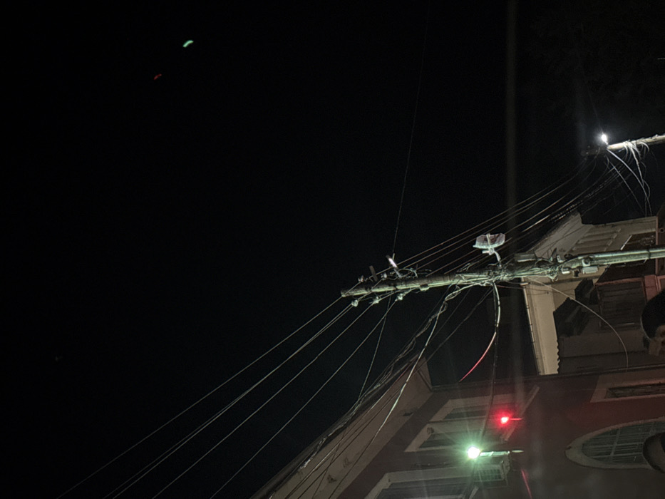 This image depicts a nighttime urban street scene in Bazan, Bulgaria. The environment is dark, illuminated by artificial light sources.
A prominent wooden utility pole stands centrally, heavily laden with numerous grey electrical and communication wires stretching in various directions across the frame. Multiple insulators and fixtures are attached to the pole, along with a small, white rectangular object secured mid-height.
To the left, a multi-story building with a reddish-brown facade is partially visible. It features at least one window with an arched top and a white cornice below the roofline. A bright green exterior light is mounted on this building, and directly below it, a distinct red light source is also present, possibly part of a traffic signal or architectural lighting.
To the far right, a modern street light with a bright white-green glow illuminates the lower right portion of the scene. In the upper right quadrant of the dark sky, two faint, small light sources are visible: one red and one green.
At the extreme bottom edge of the frame, indistinct dark shapes consistent with the tops of human heads are discernible, indicating the presence of individuals, though no specific features, actions, or interactions are visible.