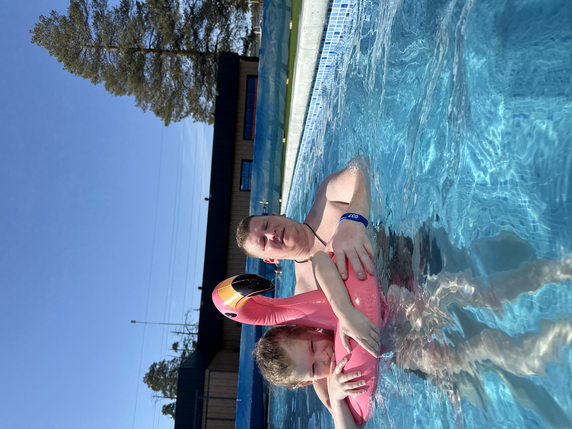 A man and a child are in a swimming pool, with the child inside a pink flamingo-shaped inflatable. The man has a blue wristband marked "037". The pool has a blue tile edge and is surrounded by a building and trees. The location is Votkinsk, Russia, as the image was taken there.
