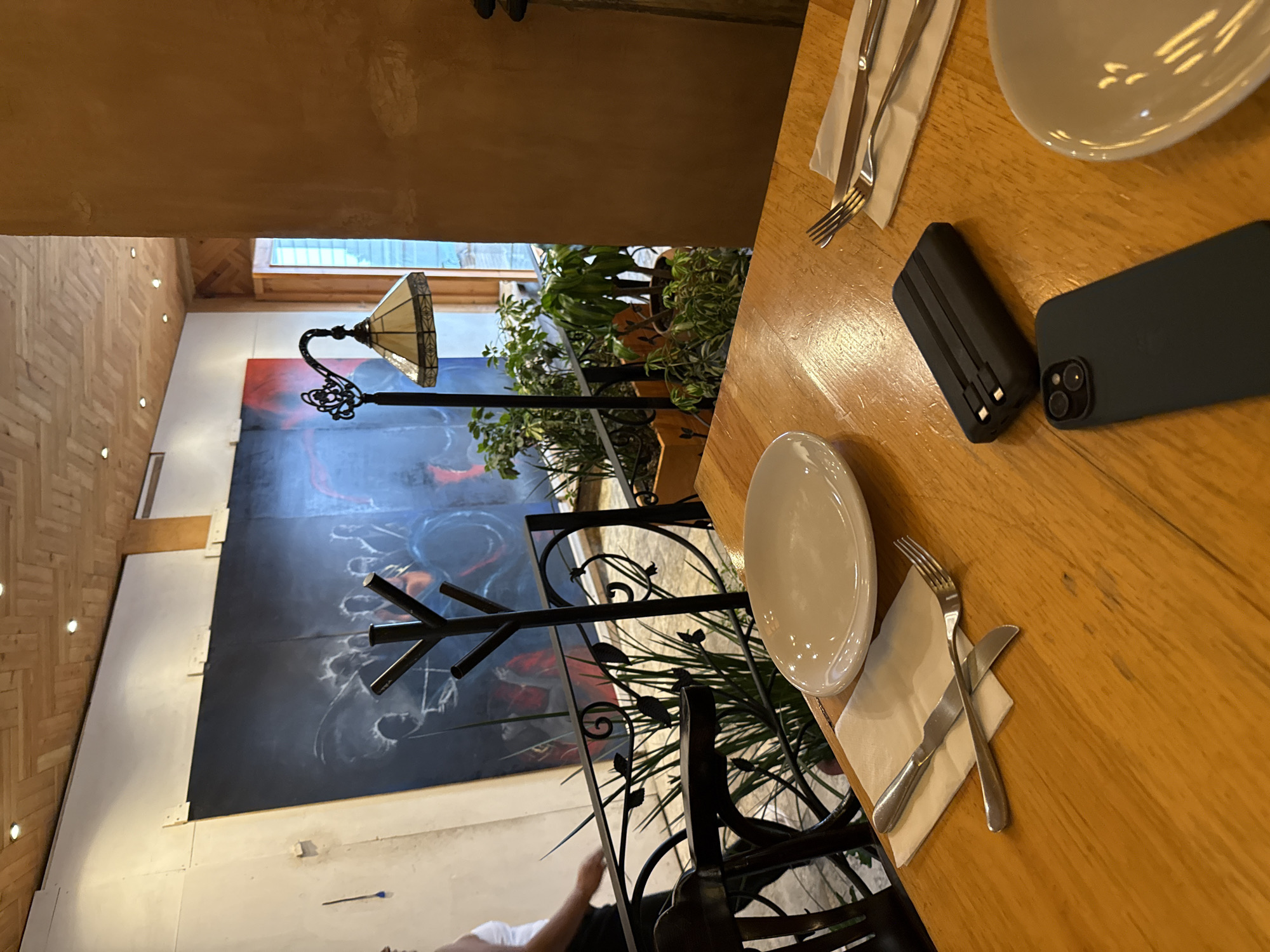 This is an indoor shot of a dining table at a restaurant in San Cristóbal, Mexico. The table is wooden and set with plates, silverware, and napkins. A black smartphone and a black portable charger are placed on the table. A decorative wrought iron railing and plants are partially visible near the table. In the background, there's a lamp and a large painting on the wall. A glimpse of a person in a white shirt can be seen near the bottom of the frame. The overall environment appears to be a restaurant setting with an artistic and natural ambiance.
