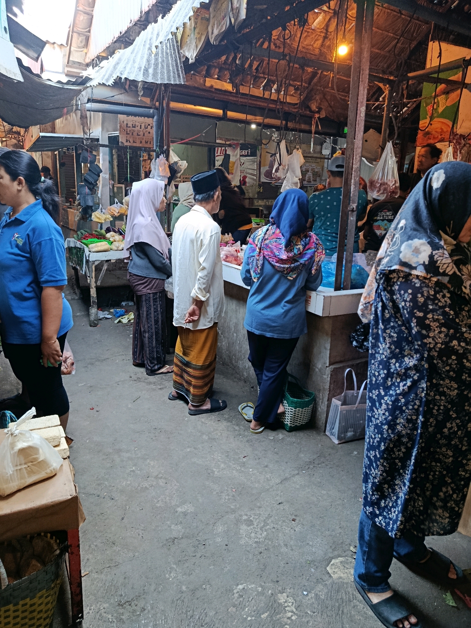 The image depicts a bustling marketplace in Bululawang, Indonesia. The scene is characterized by a covered walkway with various stalls selling goods. In the foreground, a woman wearing a blue shirt and black pants is partially visible, standing at a stall. To her right, a man in a cream-colored shirt and a sarong stands at another stall, with his hands behind his back. A woman in a pink hijab is also visible, facing away from the camera, at a stall with produce. Further back, another woman in a blue hijab and a colorful scarf is seen interacting at a stall. The stalls are laden with goods, including what appears to be produce and meat. The overall atmosphere is lively, with people engaged in shopping and transactions. The ground is paved with concrete, and the roofing appears to be made of corrugated metal and fabric. The lighting suggests it is either early morning or late afternoon.
