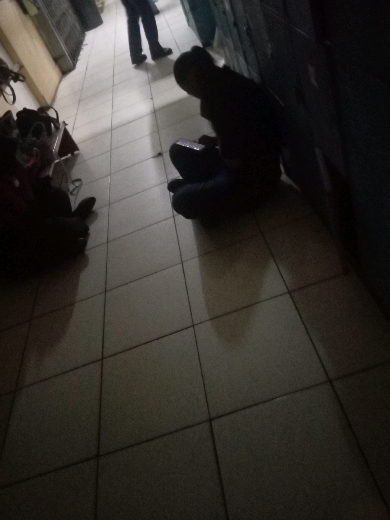 The image depicts a dimly lit hallway with tiled flooring. In the foreground, a person is seated on the floor, appearing to interact with an object on their lap. To the left, a collection of bags and a stroller are visible, suggesting the presence of individuals with luggage. In the background, another person is standing. The overall scene suggests a waiting area or a transit point. The location is identified as Batam, Indonesia, specifically within the ferry terminal building of Batam Centre. The environment appears to be an indoor public space with a functional, utilitarian design.