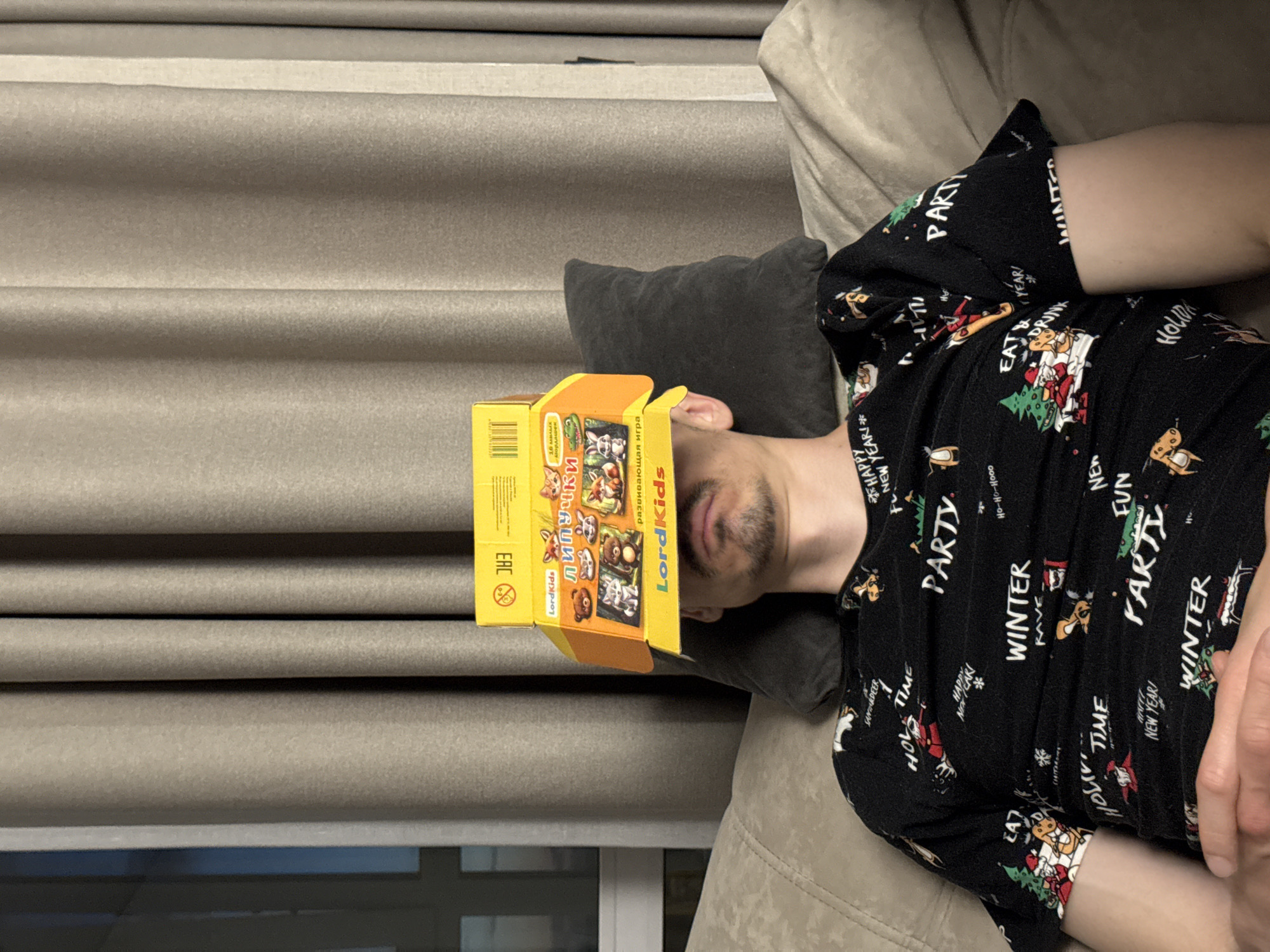 The image, originally oriented vertically and rotated right, depicts a person lying relaxed on a light-colored couch or sofa indoors. The person's head rests on a dark grey pillow, and their face is partially obscured by a small, yellow and orange cardboard box placed over their eyes and forehead. Visible facial features include light stubble on the chin and upper lip.
The individual is wearing a black long-sleeved shirt adorned with a festive, holiday-themed pattern featuring Santa Claus, reindeer, Christmas trees, presents, and snowflakes. Various words and phrases are printed on the shirt, including "PARTY", "WINTER", "RAVE", "Ho-Ho-Hooo", "HOLIDAY TIME", "EAT & DRINK", "HAPPY NEW YEAR!", "FUN", and "REINDEER".
The box covering the person's face is a product from "LordKids" and is labeled in Russian as "ЛИПУЧКИ" (Velcro/Sticky), an "развивающая игра" (developmental game) featuring "10 милых Мордашек" (10 cute faces/mugs) with cartoon animals like a fox, bear, and alligator. It also displays a barcode and an "EAC" mark.
In the background, light brown or taupe colored curtains with horizontal pleats are visible, suggesting Roman blinds. A dark window or opening is partially seen at the bottom left, behind the curtains. The overall setting appears to be a home environment, with dim lighting. The person's posture suggests they are resting or sleeping. The context given is Izhevsk, Russia.