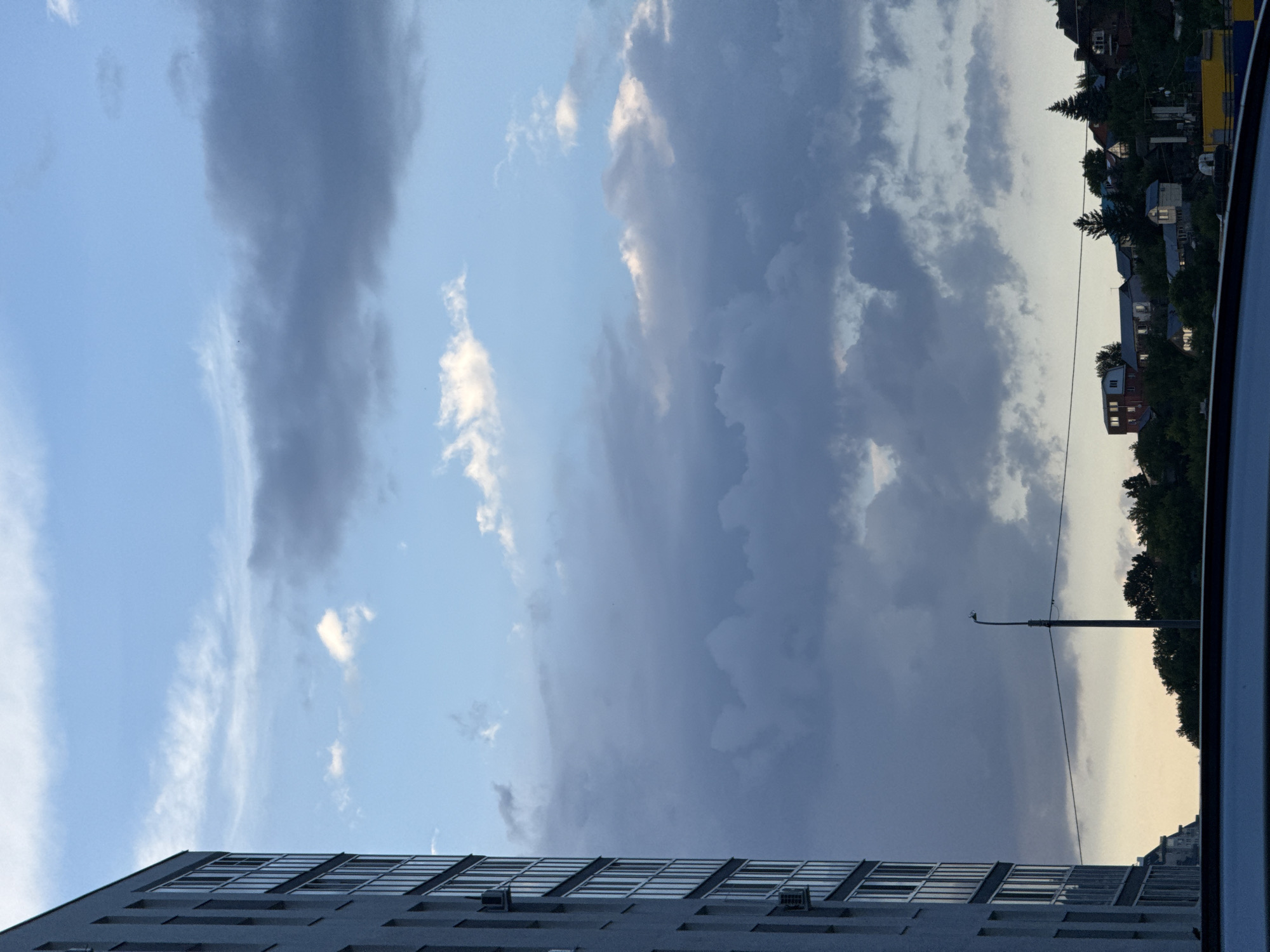 A portion of a modern apartment building is visible on the left side of the frame, characterized by its gray facade and numerous windows. The building occupies the left third of the image. In the background, a dense cluster of houses is situated on a hillside, interspersed with trees. A streetlight stands prominently in the mid-ground, with power lines stretching across the scene. The sky is filled with a mix of light and dark clouds, suggesting a dramatic or changing weather condition. The lower edge of the image shows the dark, curved shape of a vehicle's roof or hood. The overall setting appears to be a residential area within a city, under a cloudy sky.