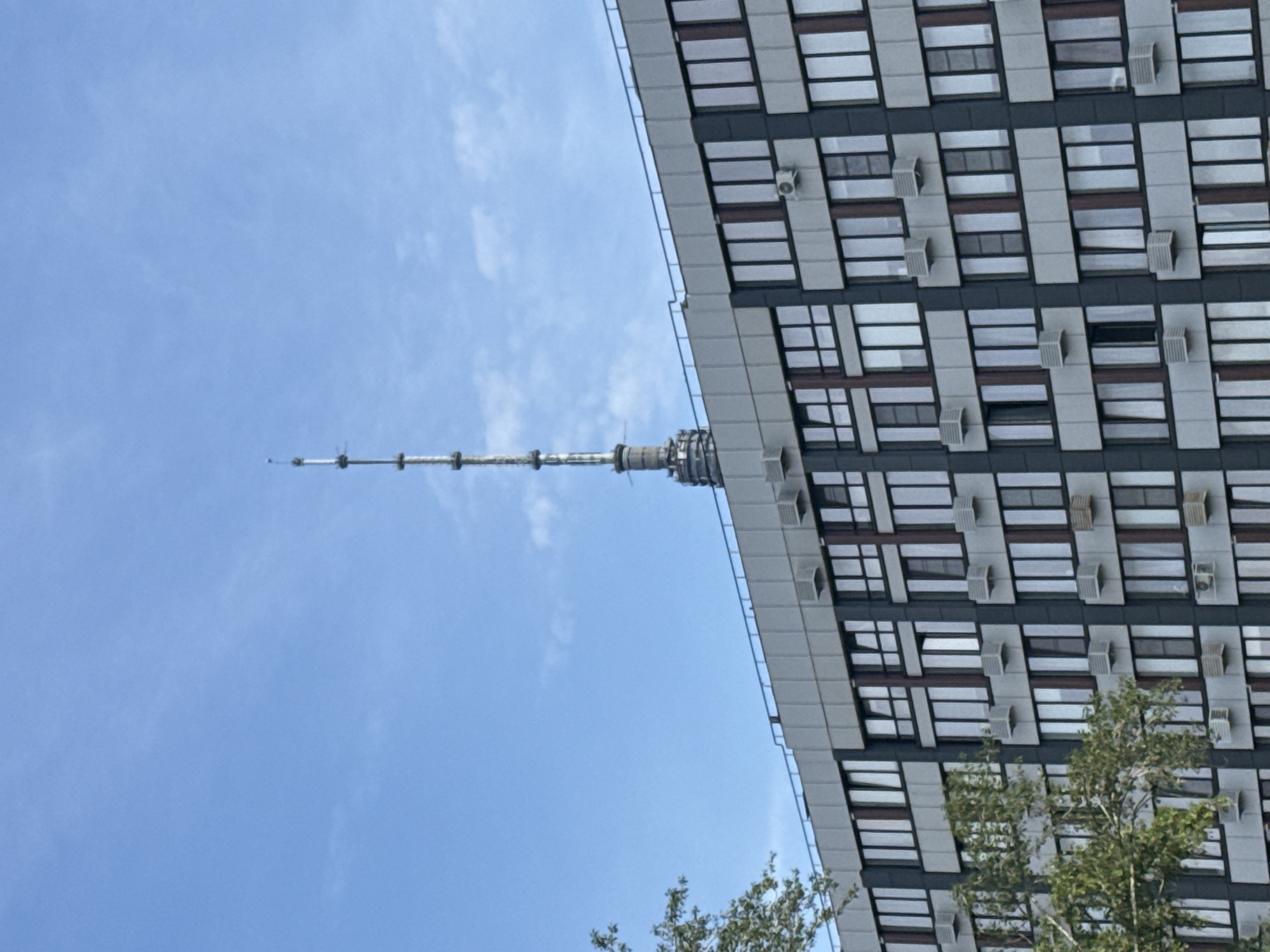 The image shows a telecommunication tower in Moscow, Russia, rising above a modern multi-story building. The tower, which appears to be the Ostankino Tower, has a slender, segmented structure with antennas at its apex. The building below is characterized by a repeating pattern of dark-framed windows and light-colored facade panels, interspersed with visible air conditioning units. A tree with green foliage partially obscures the left side of the building. The sky is a clear, light blue with a few wispy clouds. The overall scene depicts a juxtaposition of urban infrastructure and natural elements under a bright sky.
